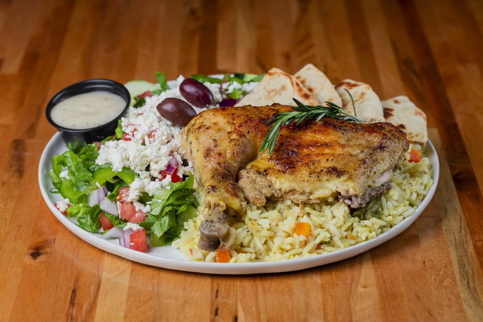 Platter of chicken leg quarter with rice, salad, pita bread,and a side of white dipping sauce on a white oval plate on wooden table.