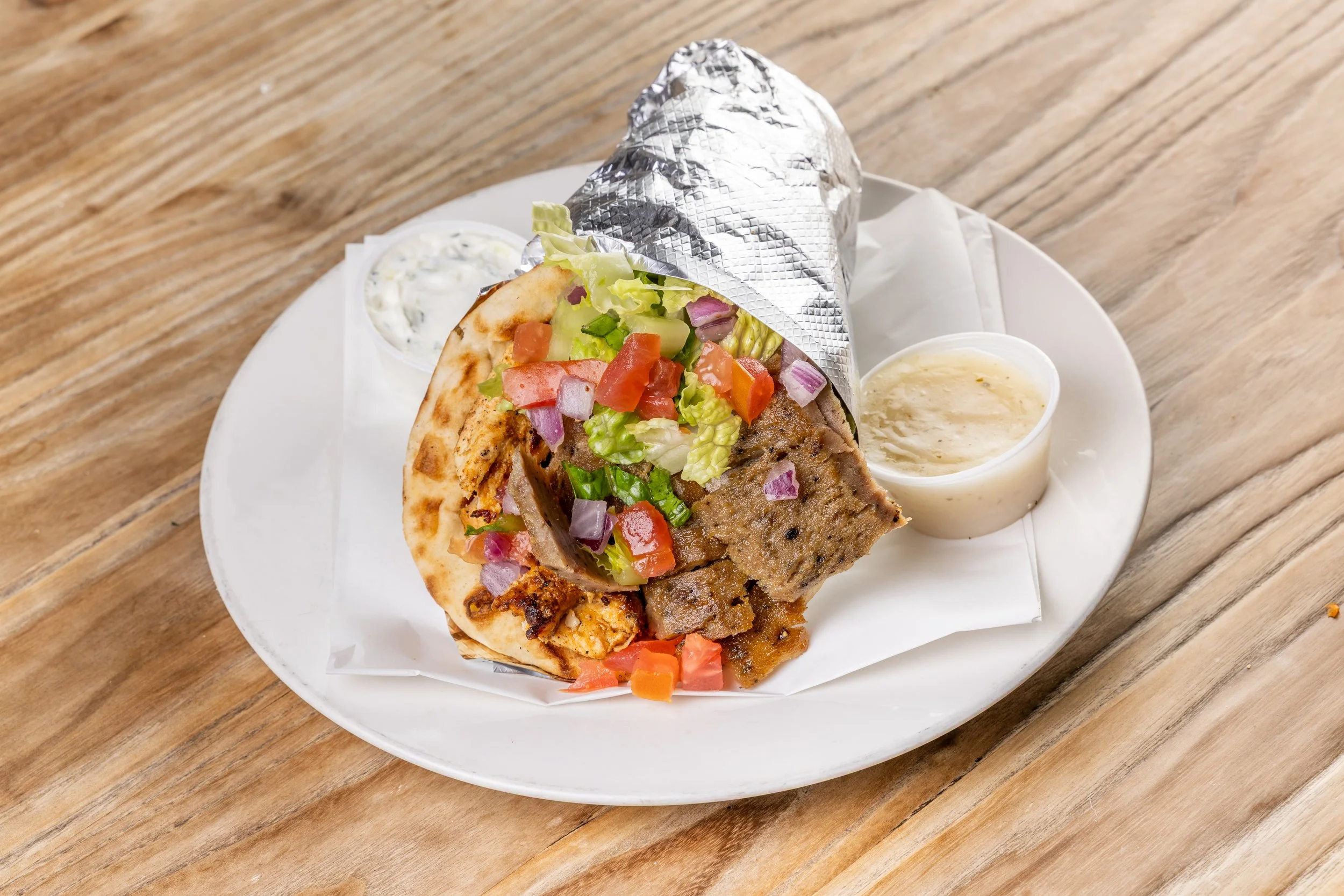 Beef/lamb gyro pita wrapped in foil with lettuce, tomato, onion, served with two side sauces on a white plate on a wooden table.