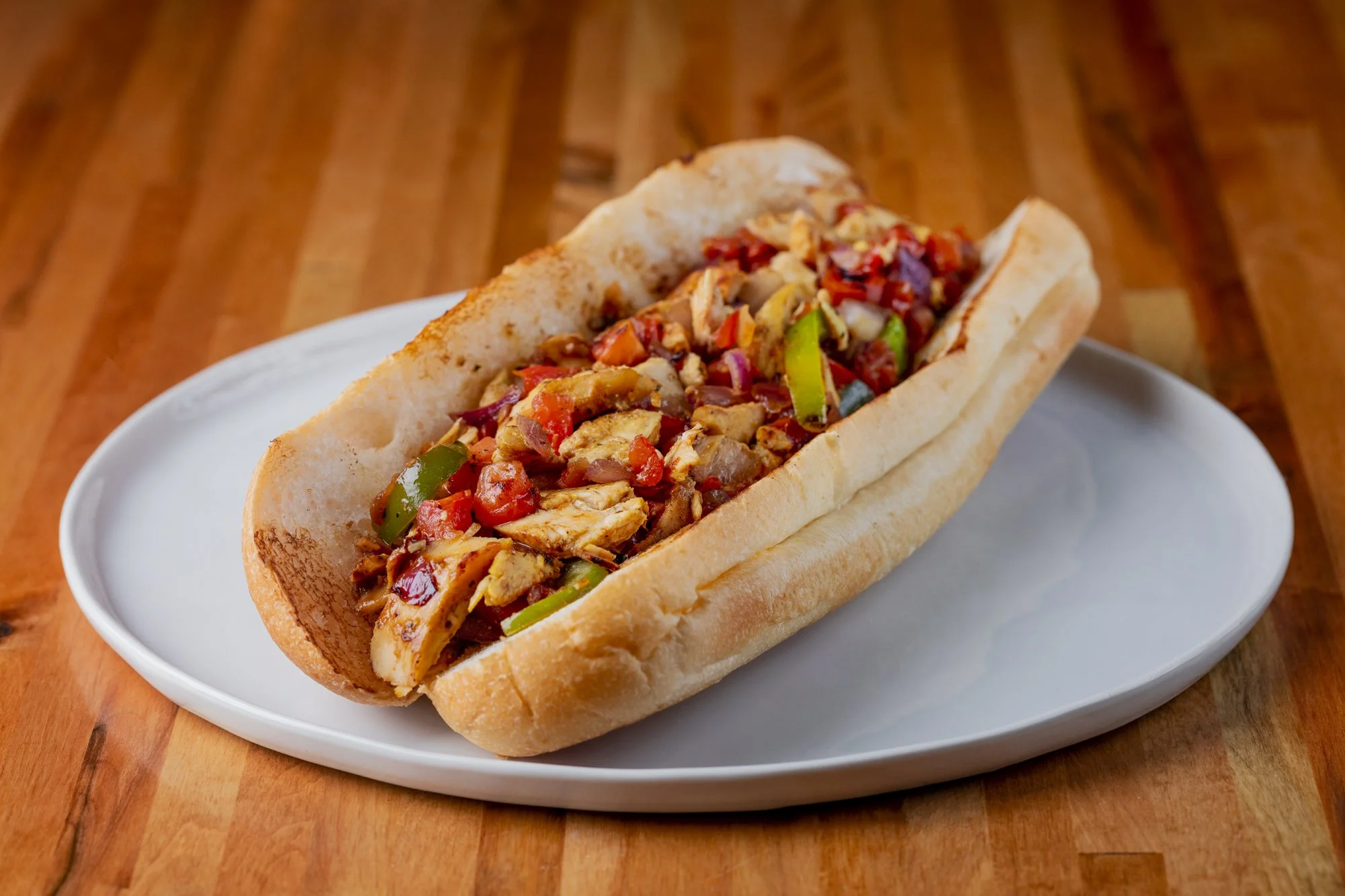 chicken philly served in a hoagie roll on a white plate with grilled peppers, tomatoes, and onions, on a wooden table.