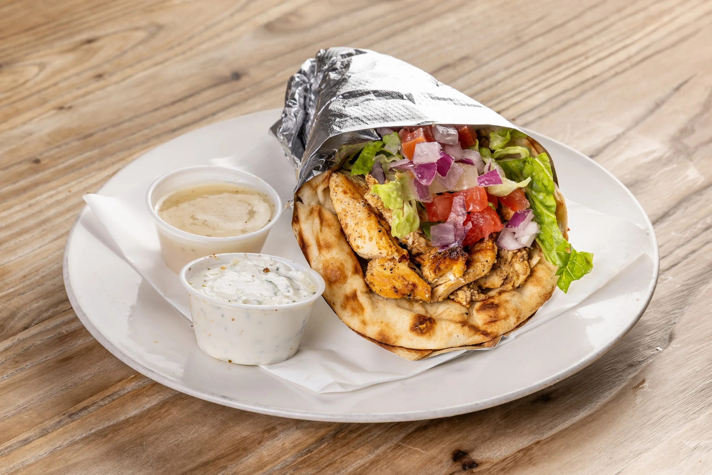 Chicken gyro pita wrapped with chopped vegetables, served with two sauces on a white plate.
