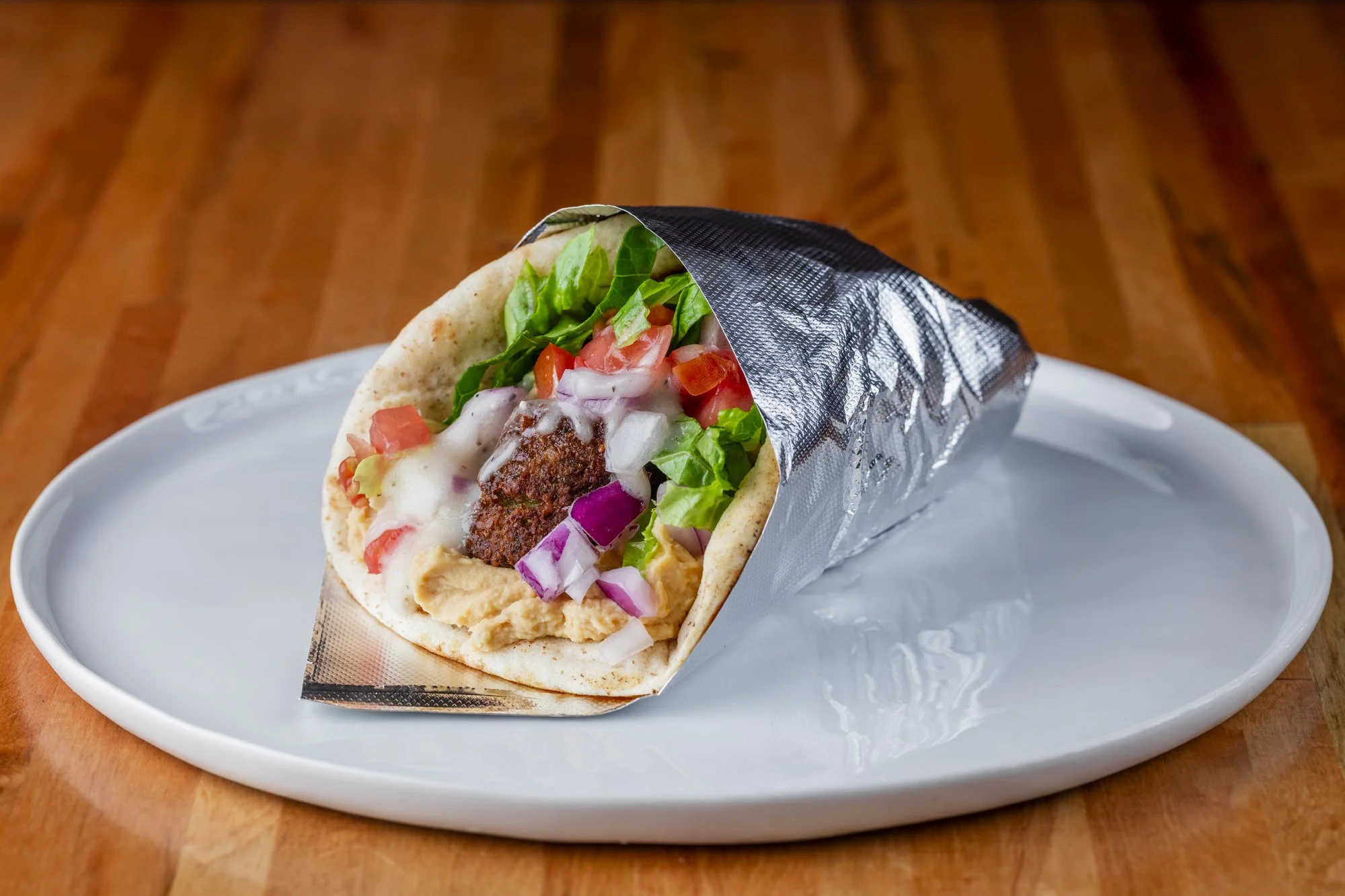 A falafel pita wrapped with lettuce, tomatoes, onions, hummus, and Greek dressing on a white plate.