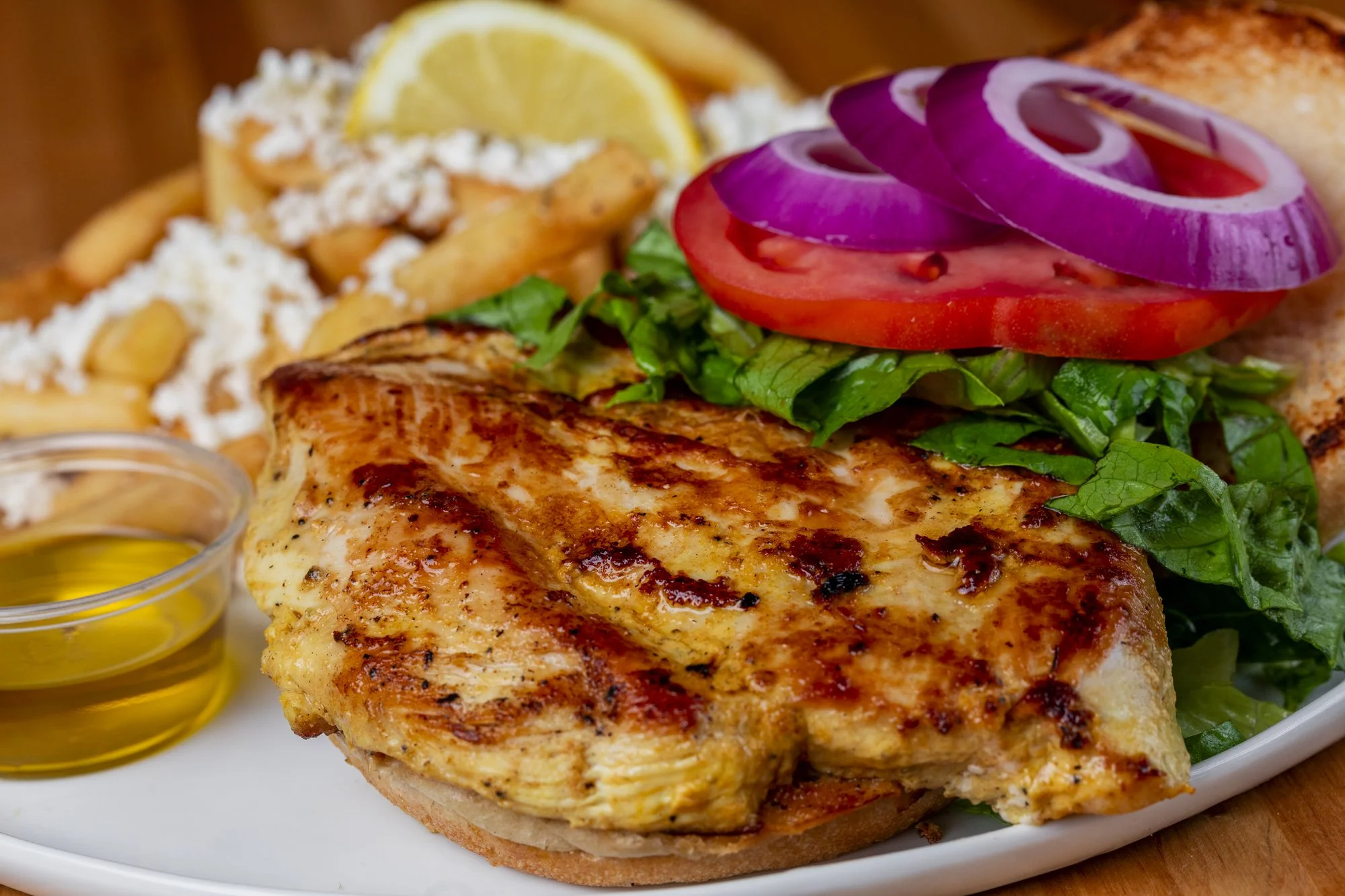 Grilled chicken breast with fresh tomato, red onion, and lettuce ona kaiser roll, served with lemon wedge, French fries with crumbled cheese, and a small cup of olive oil