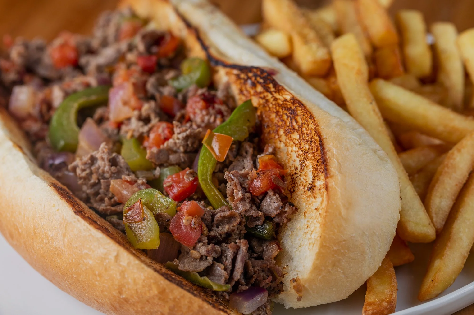Close-up of takeout Philly cheesesteak sandwich with cooked chopped steak, green bell peppers, and tomatoes in a hoagie bun, served with side of French fries.