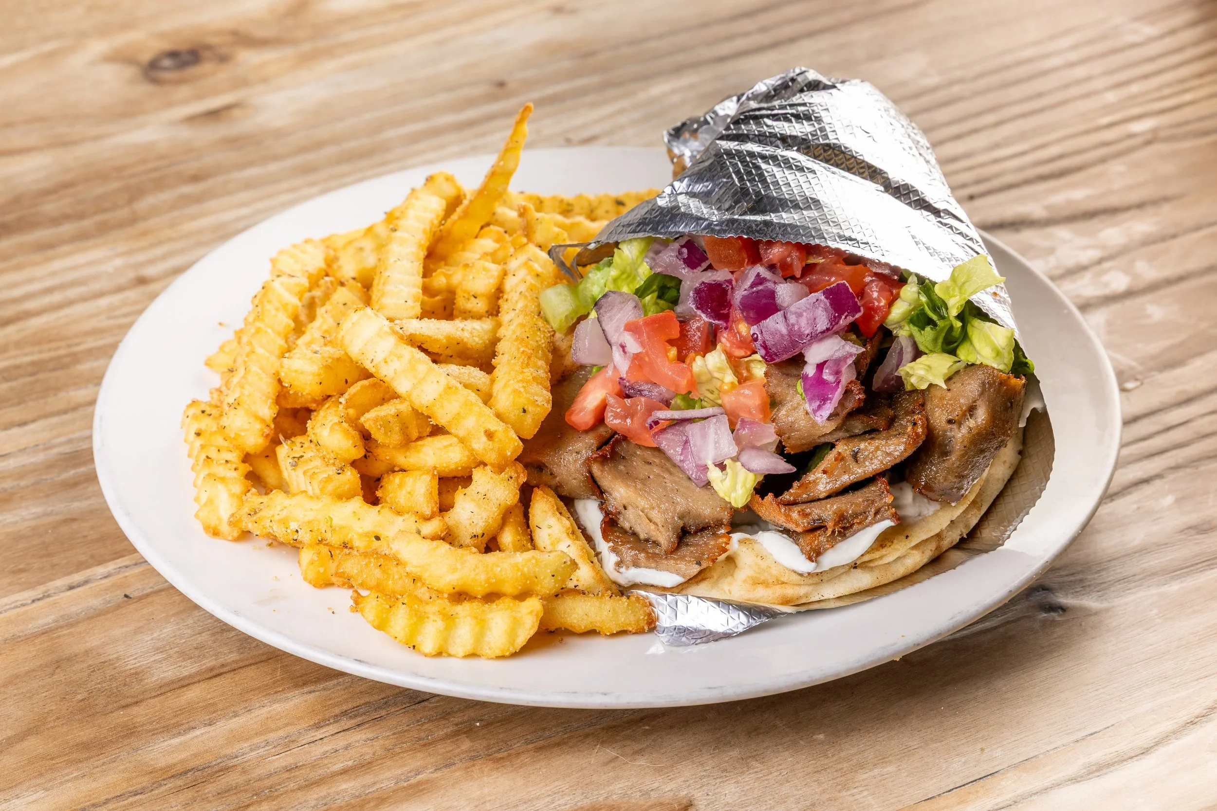 A plate of French fries served with a beef/lamb gyro pita wrapped with fresh vegetables on a wooden table.