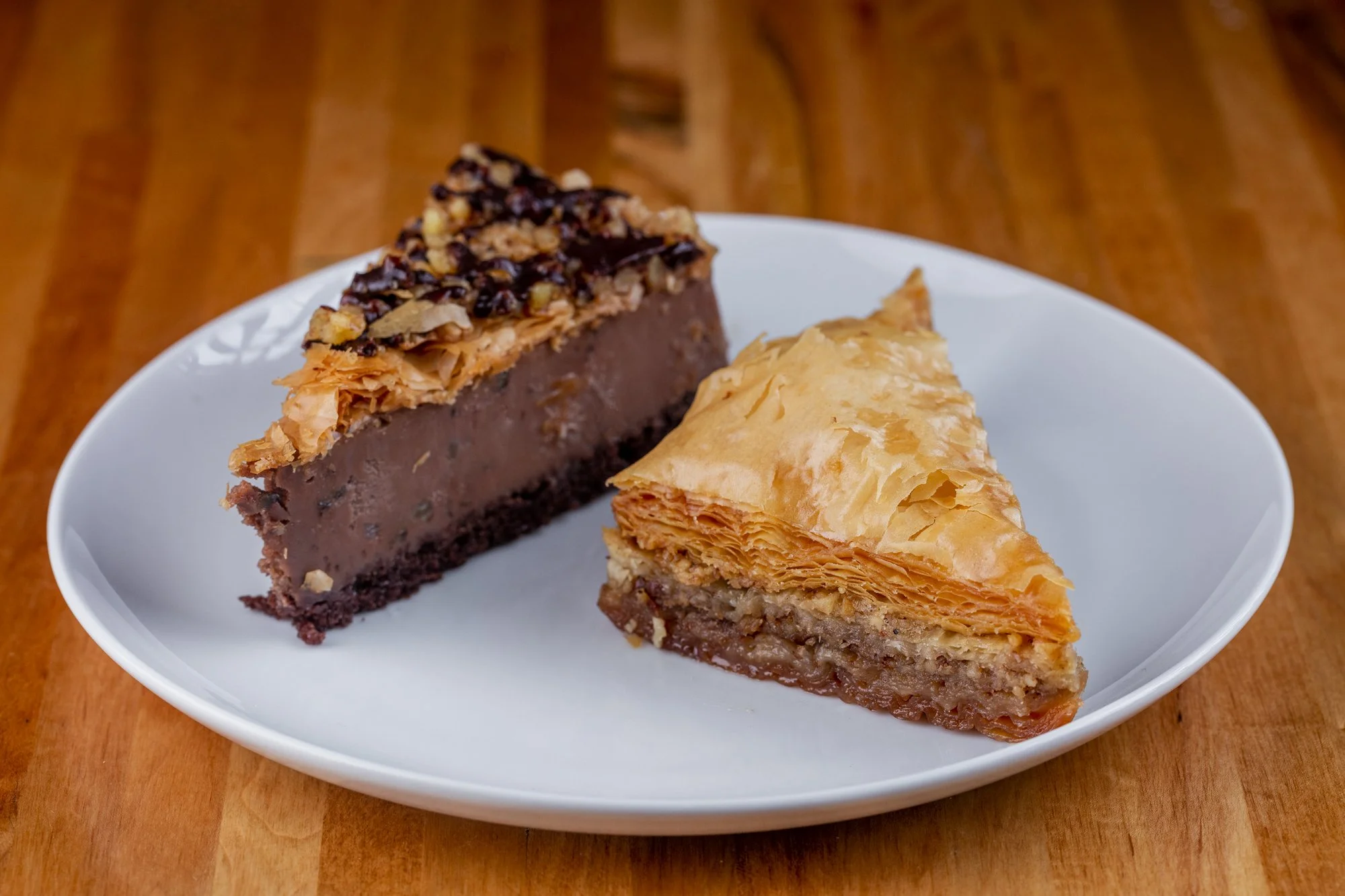 Two slices of Greek desserts on a white plate, one with chocolate filling and a flaky crust on top, the other with a golden, layered crust and nut filling inside, placed on a wooden surface.
