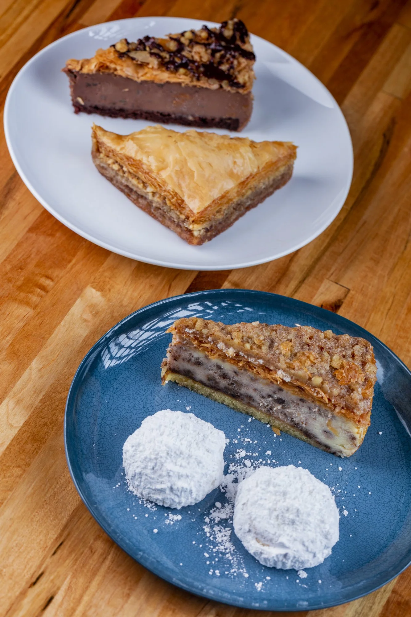 Two plates of assorted Greek pastries and cookies on a wooden table, with powdered sugar dusted on cookies