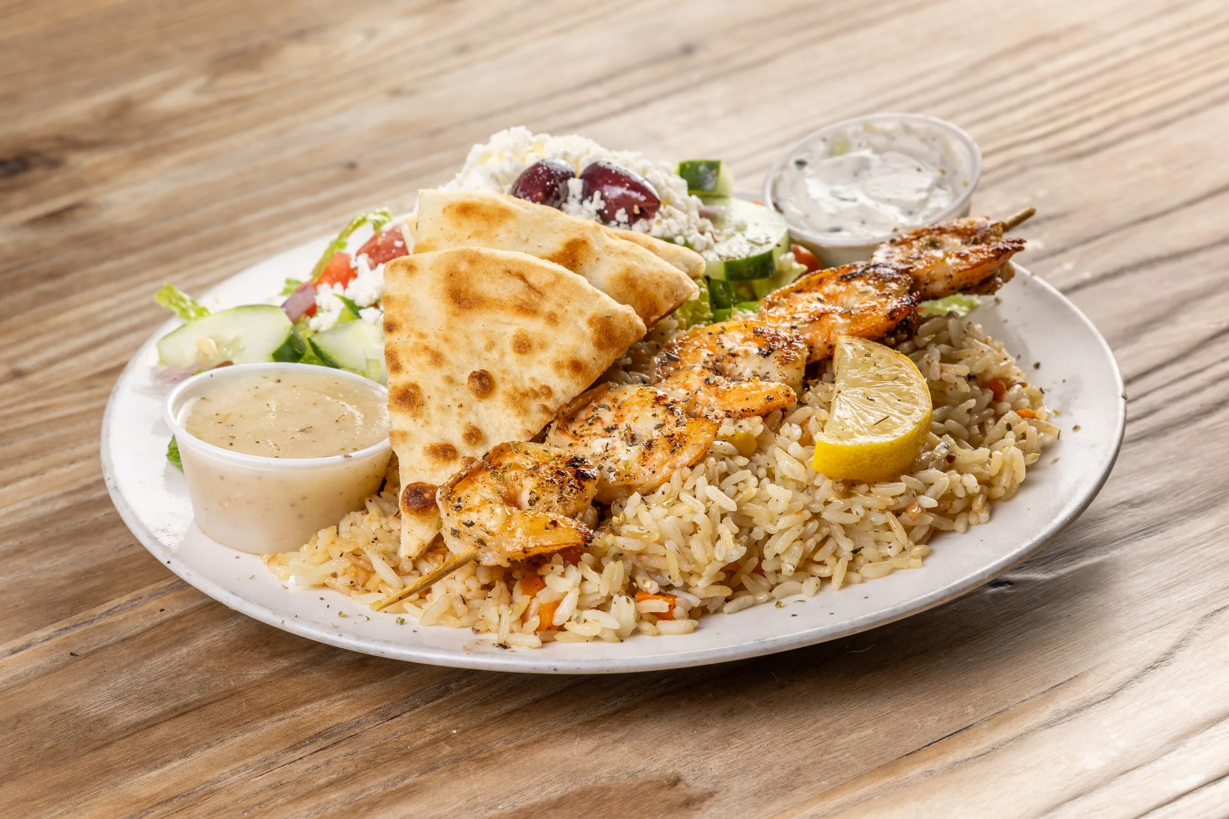 Plate of grilled shrimp on a skewer with rice, lemon wedge, pita bread, Greek salad, and two dipping sauces on a wooden table.