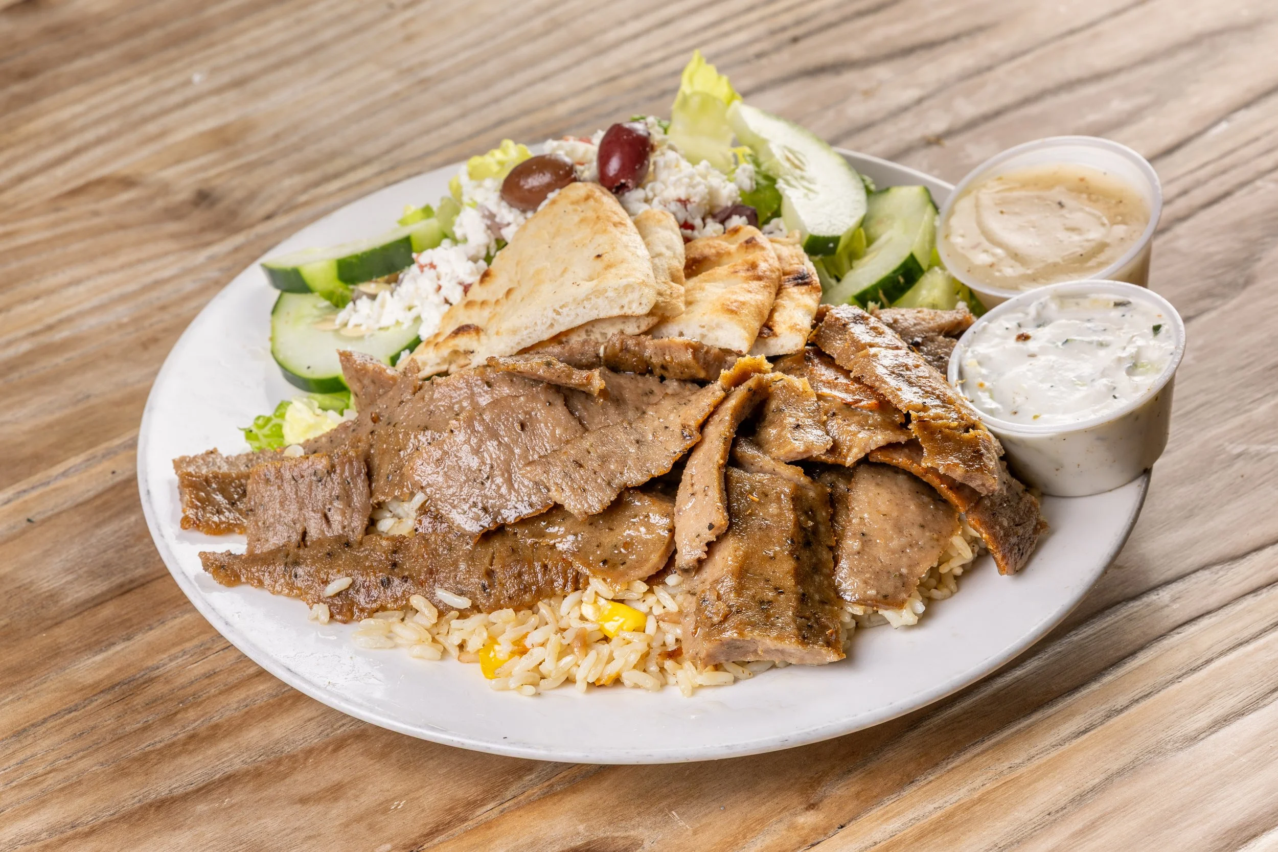 Plate of with sliced beef/lamb gyro meat, rice, Greek salad with pita bread, and two small cups of sauces on a wooden table.