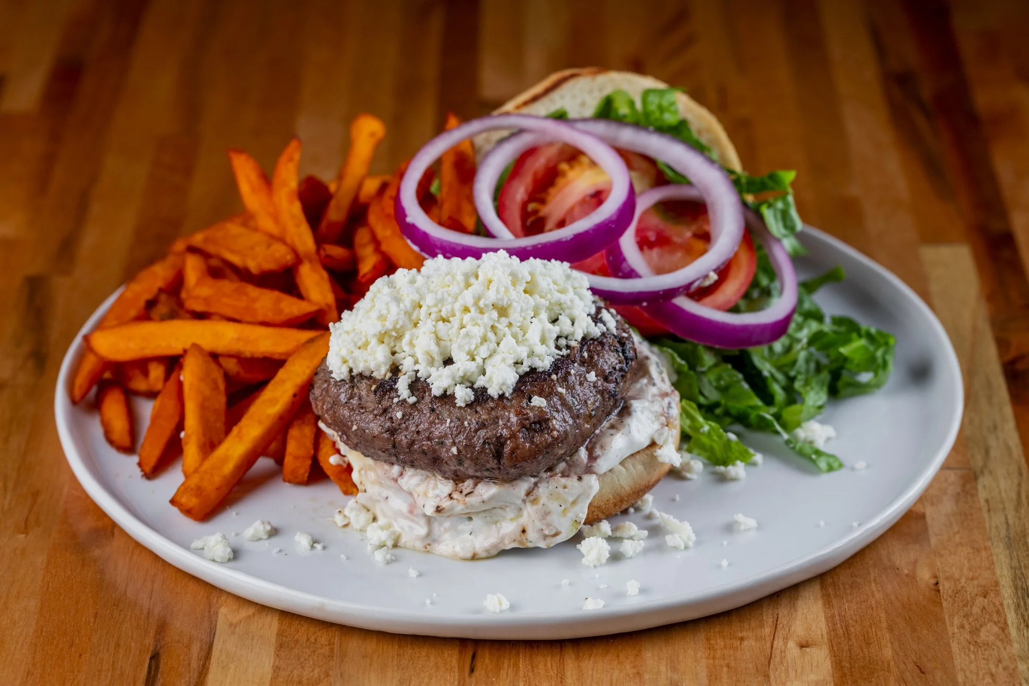 Plate with sweet potato fries, burger with grilled beef patty, shredded lettuce, sliced tomatoes, and onions on a wooden table.