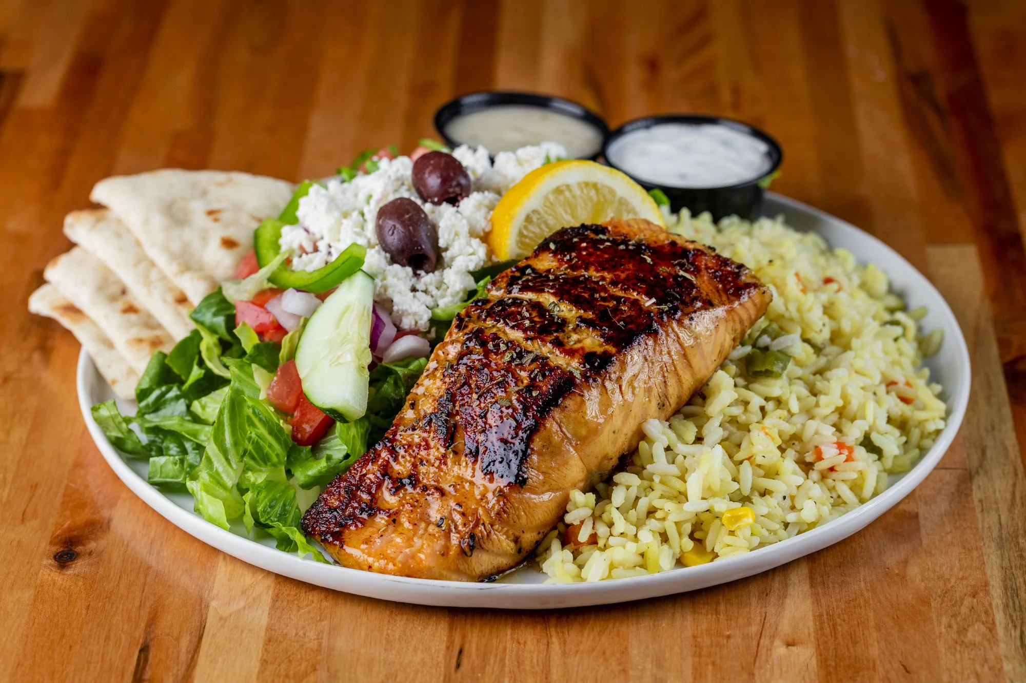 Grilled salmon with rice, Greek salad, pita bread, lemon, and dressings on a white oval plate