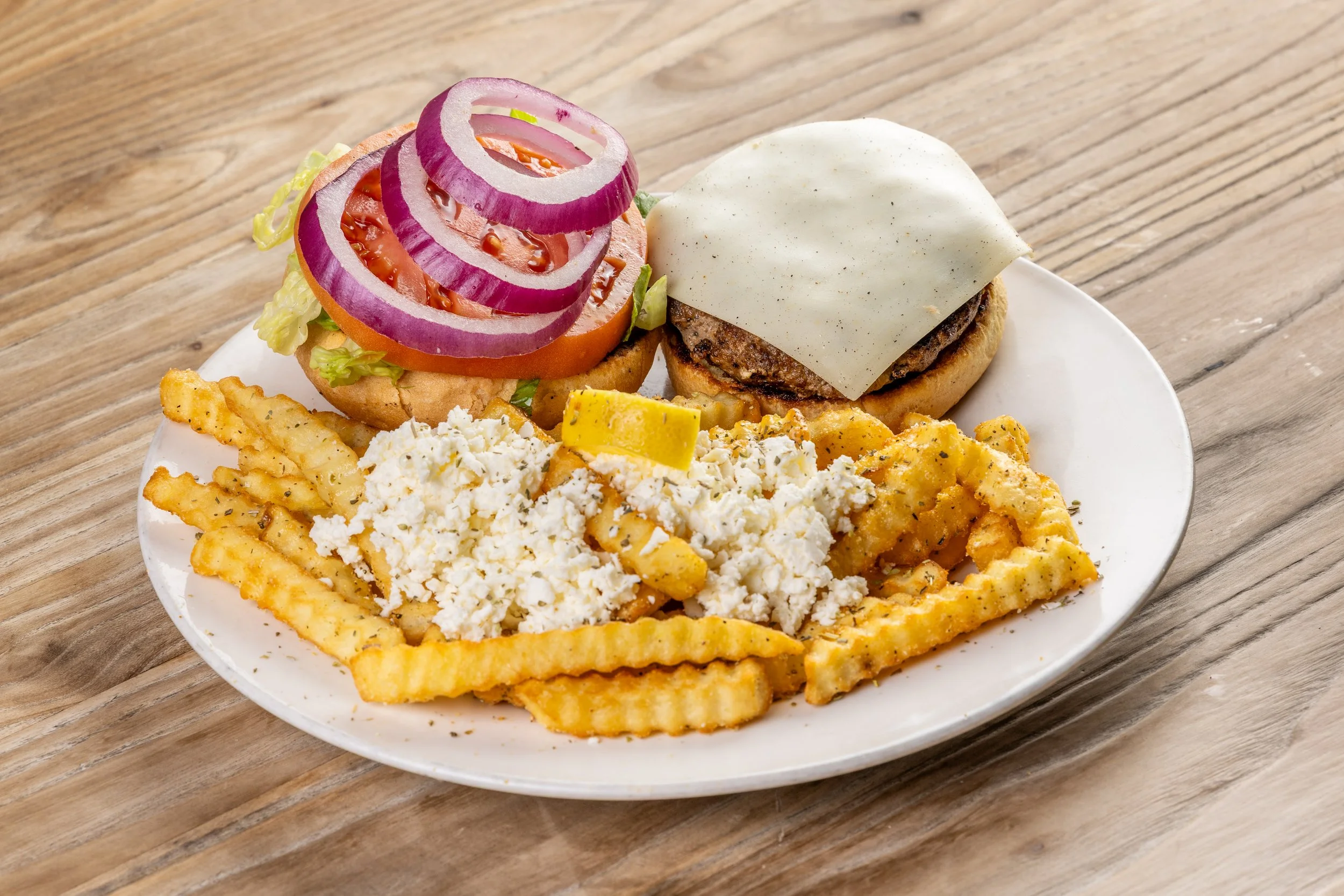 Plate of a cheeseburger with tomato, onion, lettuce, and white American cheese with a side of crinkle-cut fries topped with feta cheese crumbles, all on a wooden table.