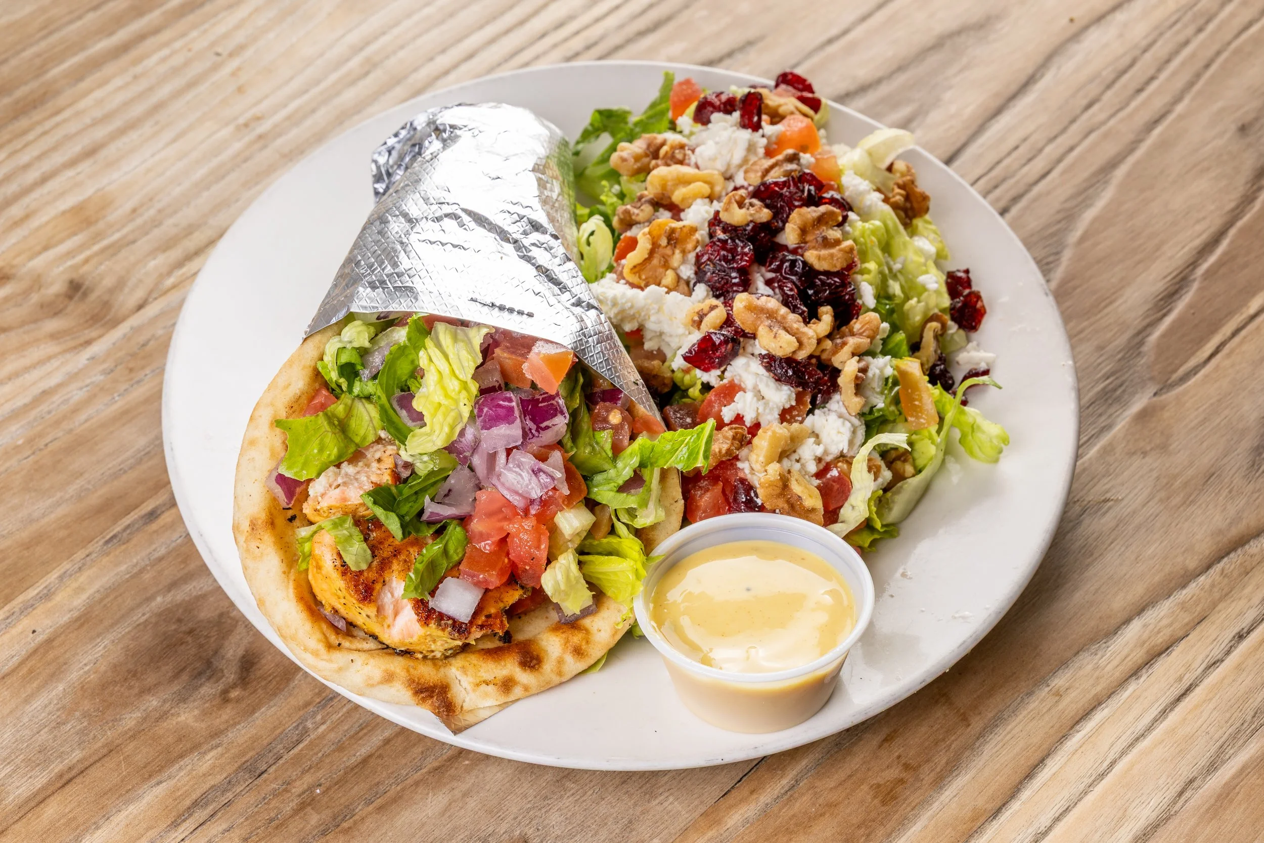 A plate with a salmon pita wrap partially covered with foil, a mixed green salad topped with feta cheese, dried cranberries, walnuts, and diced vegetables, and a small cup of creamy dressing, all on a wooden table.