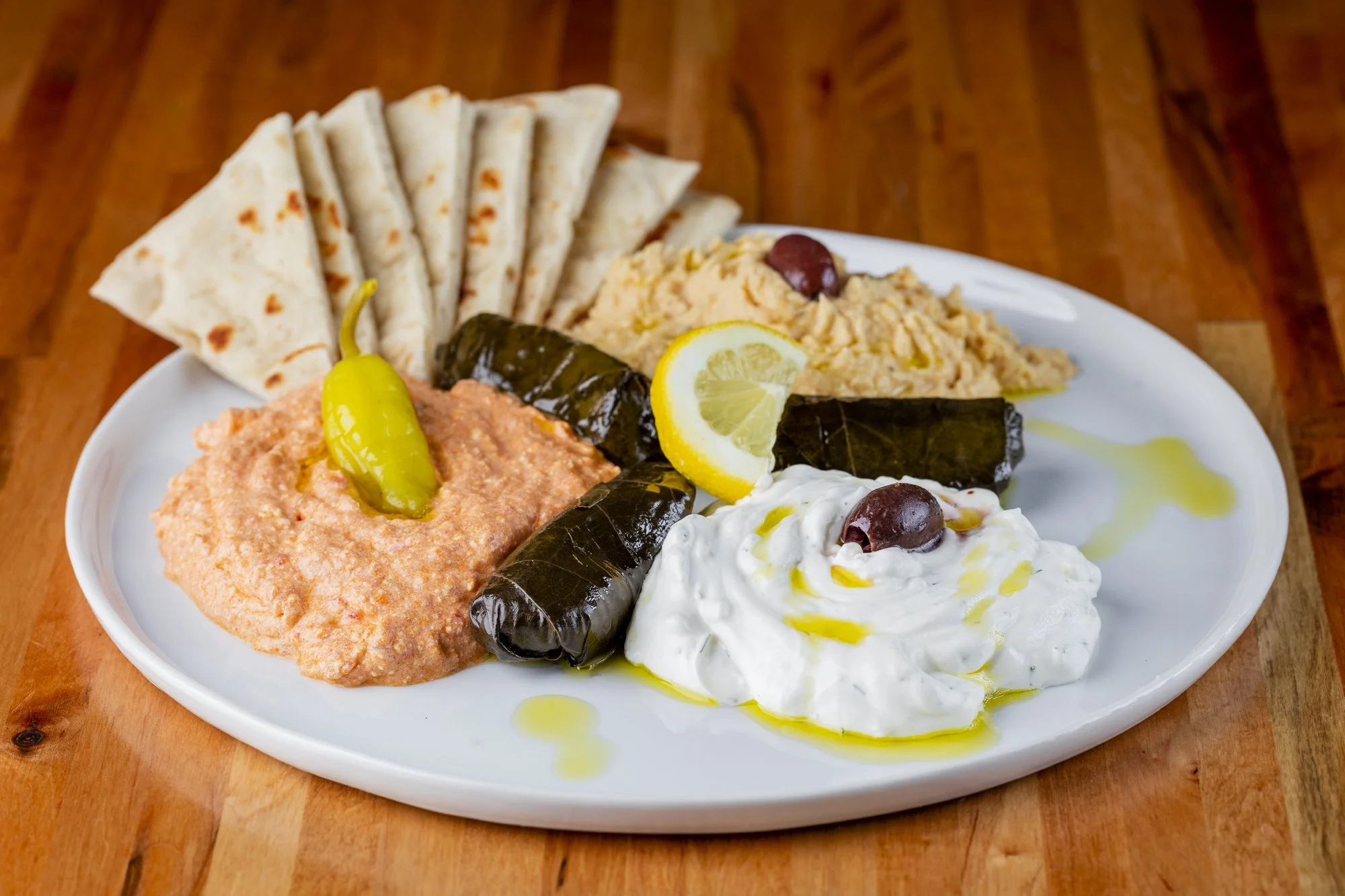 Plate of Greek appetizers including hummus topped with an olive, stuffed grape leaves, a portion of tzatziki, a dollop of spicy feta spread with pita slices, and a lemon wedge.