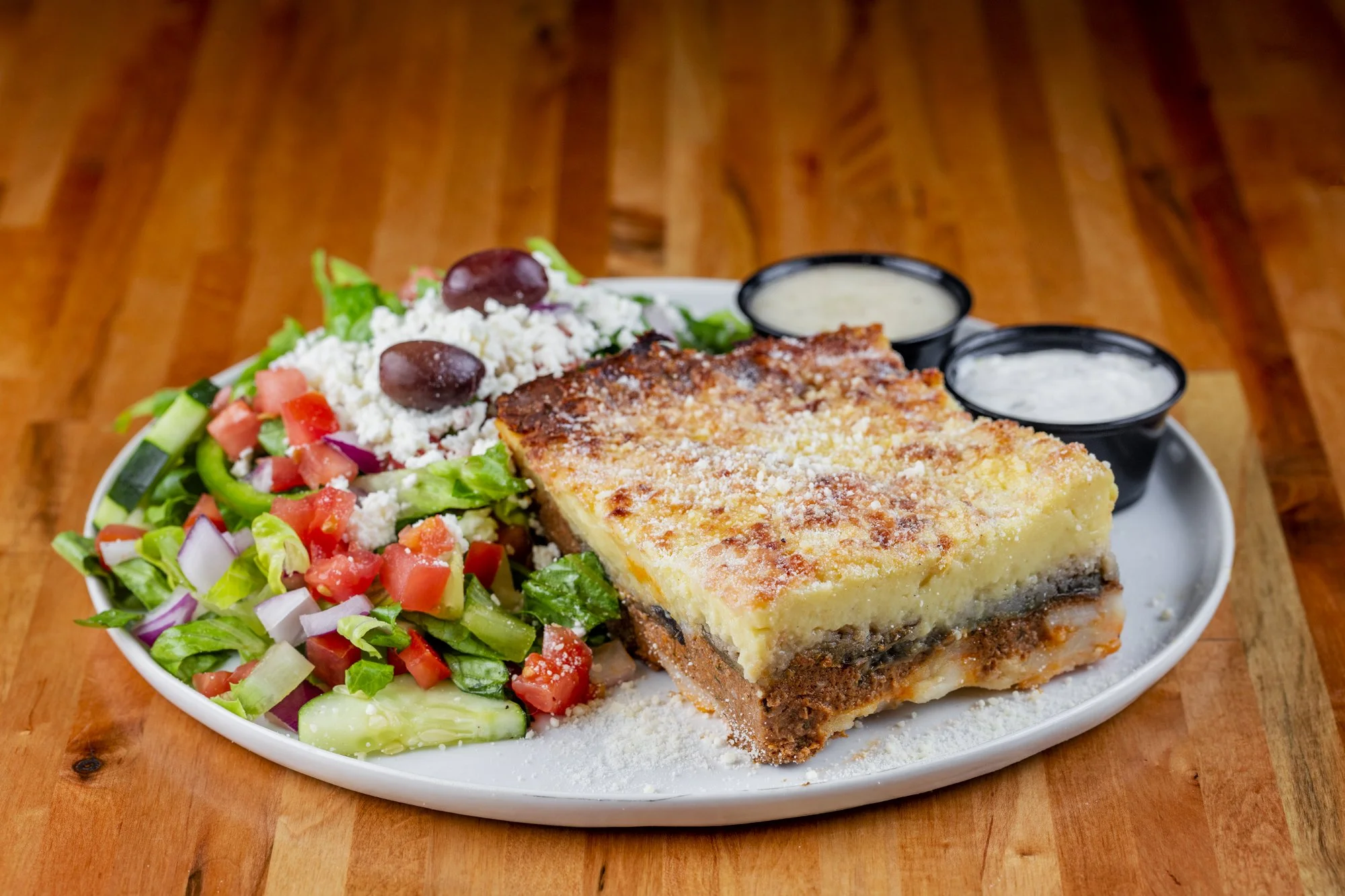 A plate of Mousaka layered with potatoes, ground beef, and bechamel sauce with a sides of Greek salad, and sauces on a wooden table.