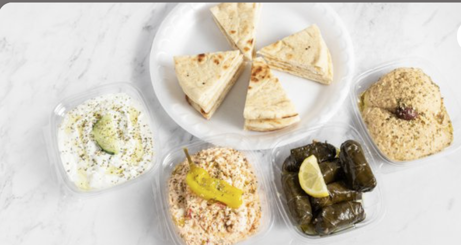 Plate with pita wedges, and four containers with Mediterranean appetizers: hummus with an olive, spicy feta dip with a pepperoncini, tzatziki with a slice of cucumber, and stuffed grape leaves.