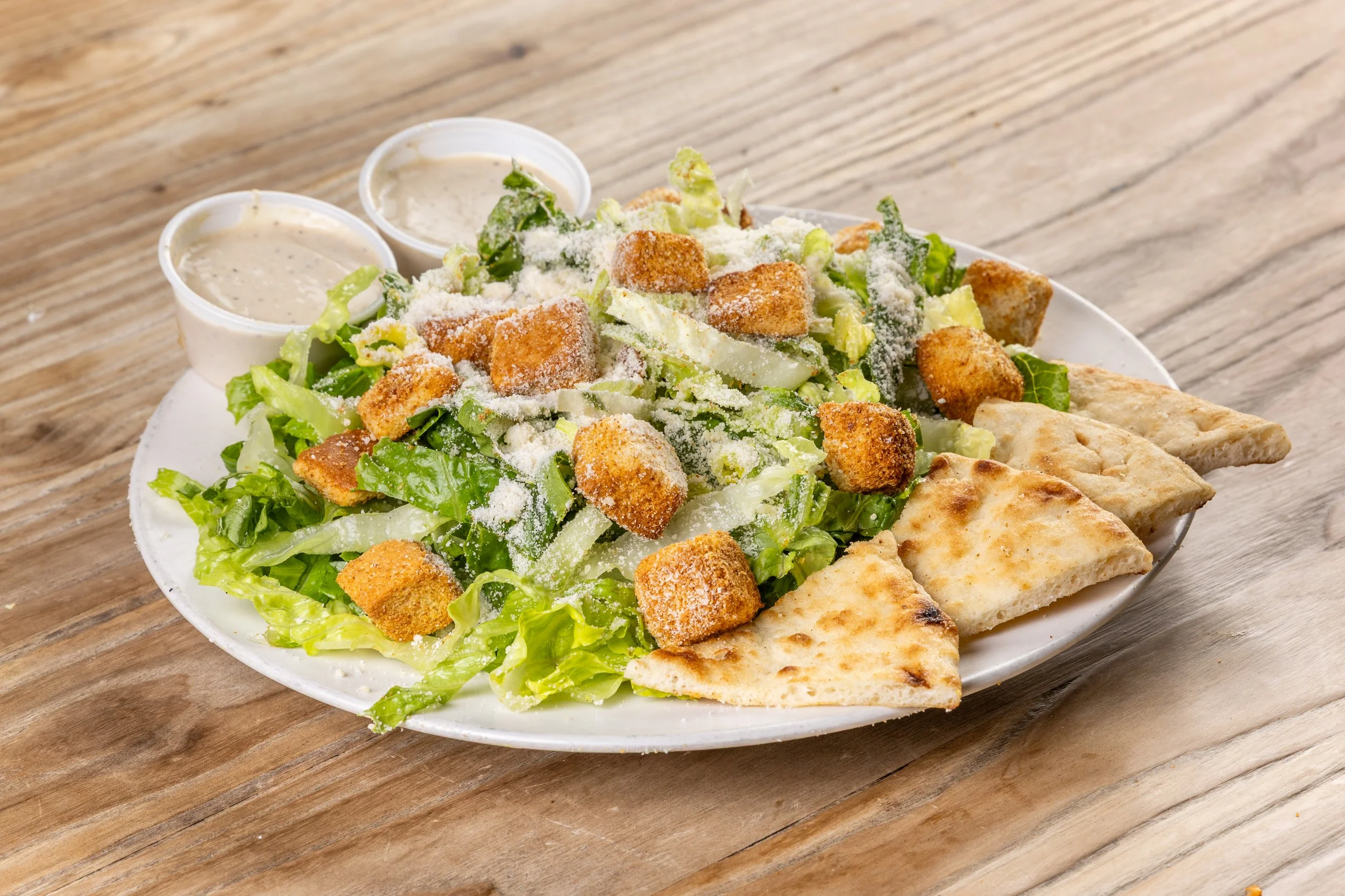 Caesar salad with romaine lettuce, croutons, Parmesan cheese, served with pita bread triangles, and two small containers of Caesar dressing, on a wooden table.