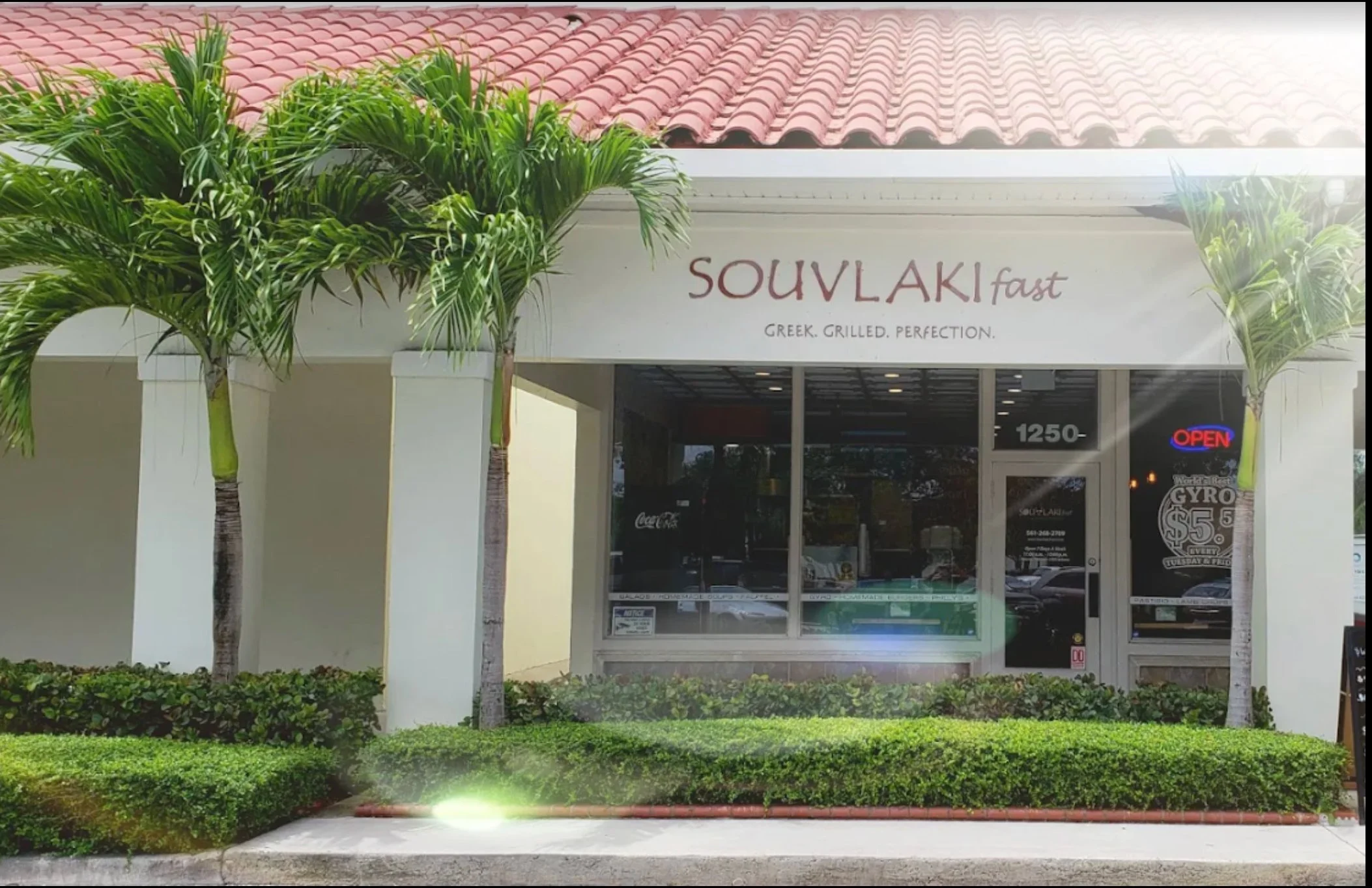 Exterior of a Greek restaurant in Royal Palm Beach named Souvlaki Fast with palm trees and well-maintained bushes outside, a large window, and a door with a neon open sign.