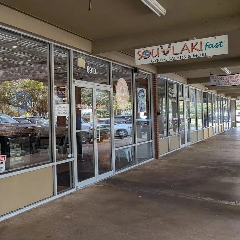 Exterior view of a Greek restaurant in Palm Beach Gardens called Souvlaki Fast offering gyros, salads, and more, with glass windows and doors, and parking lot outside.