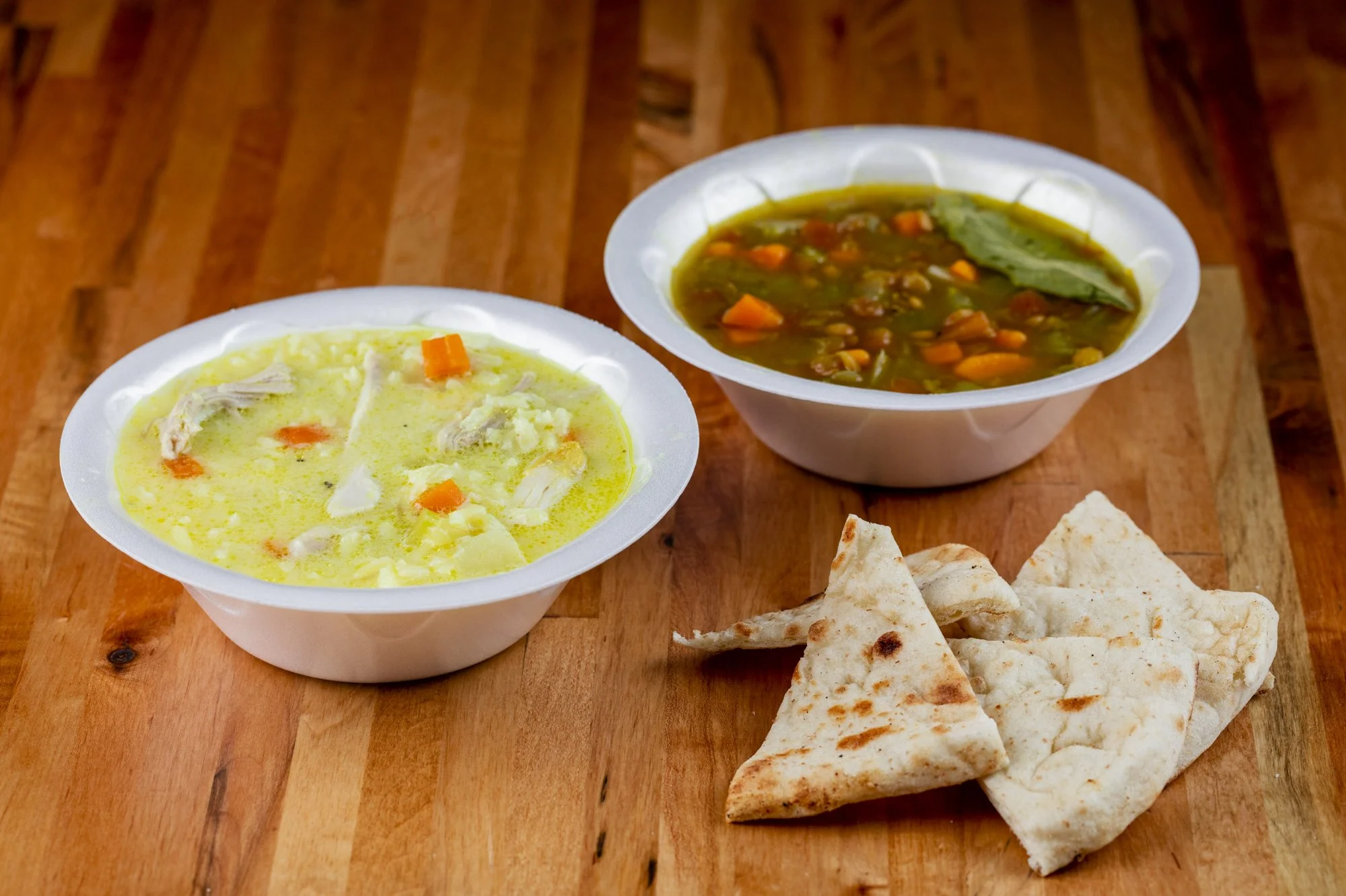 Two bowls of soup, one with lemon chicken soup and the other with lentil soup, served with pieces of pita bread on a wooden surface.