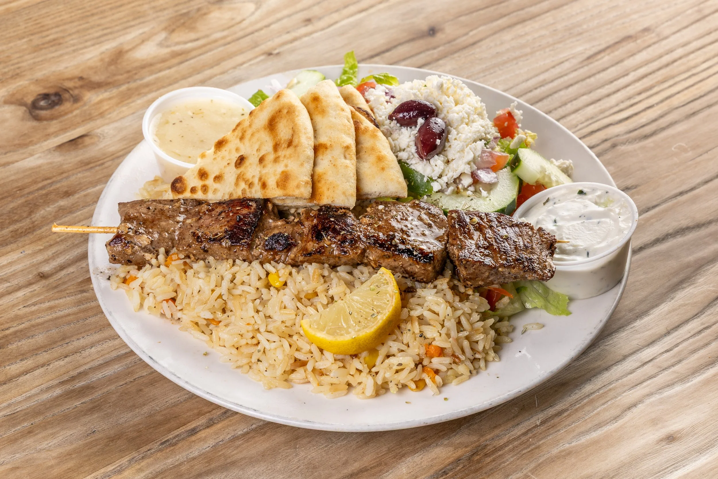 A plate of a beef souvlaki with rice, pita bread, Greek salad, two small containers of sauce, and a lemon wedge.