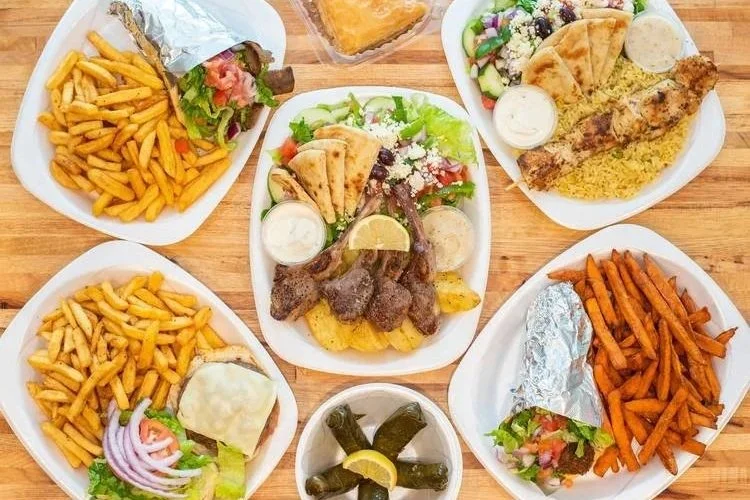 Plates of French fries, grilled lamb chops, pita wraps, souvlaki, cheeseburger, saladss and stuffed grape leaves on a wooden table.