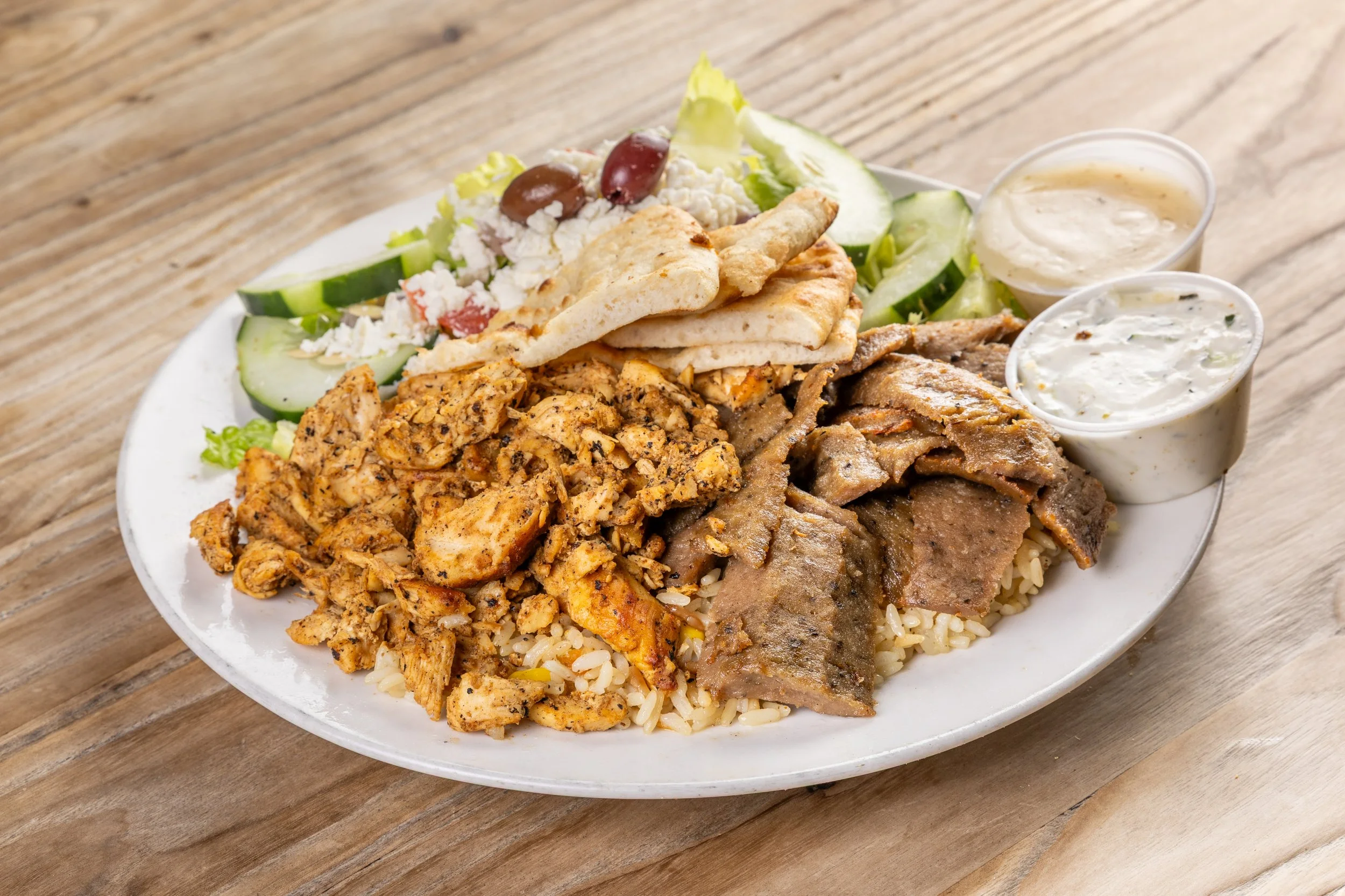 A plate of with chicken gyro and beef/lamb gyro meat, rice, Greek salad, pita bread, and two kinds of sauce.