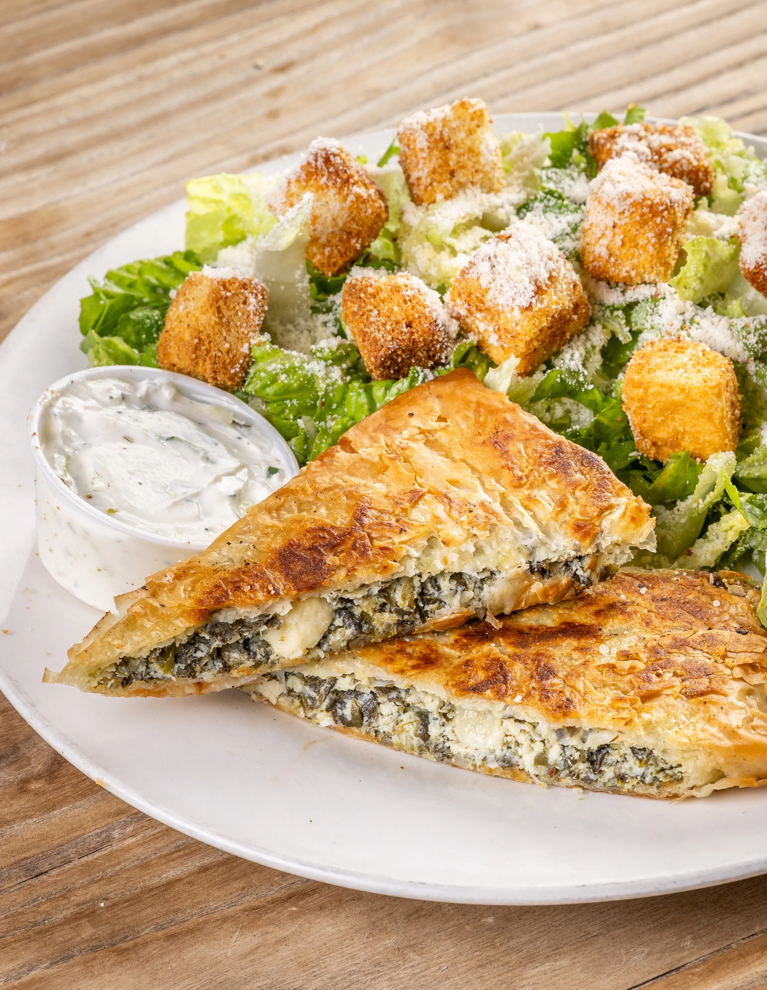 A plate with a piece of spinach pie cut in half and a caesar salad topped with croutons, grated cheese, and shredded lettuce, with a side of creamy dressing.