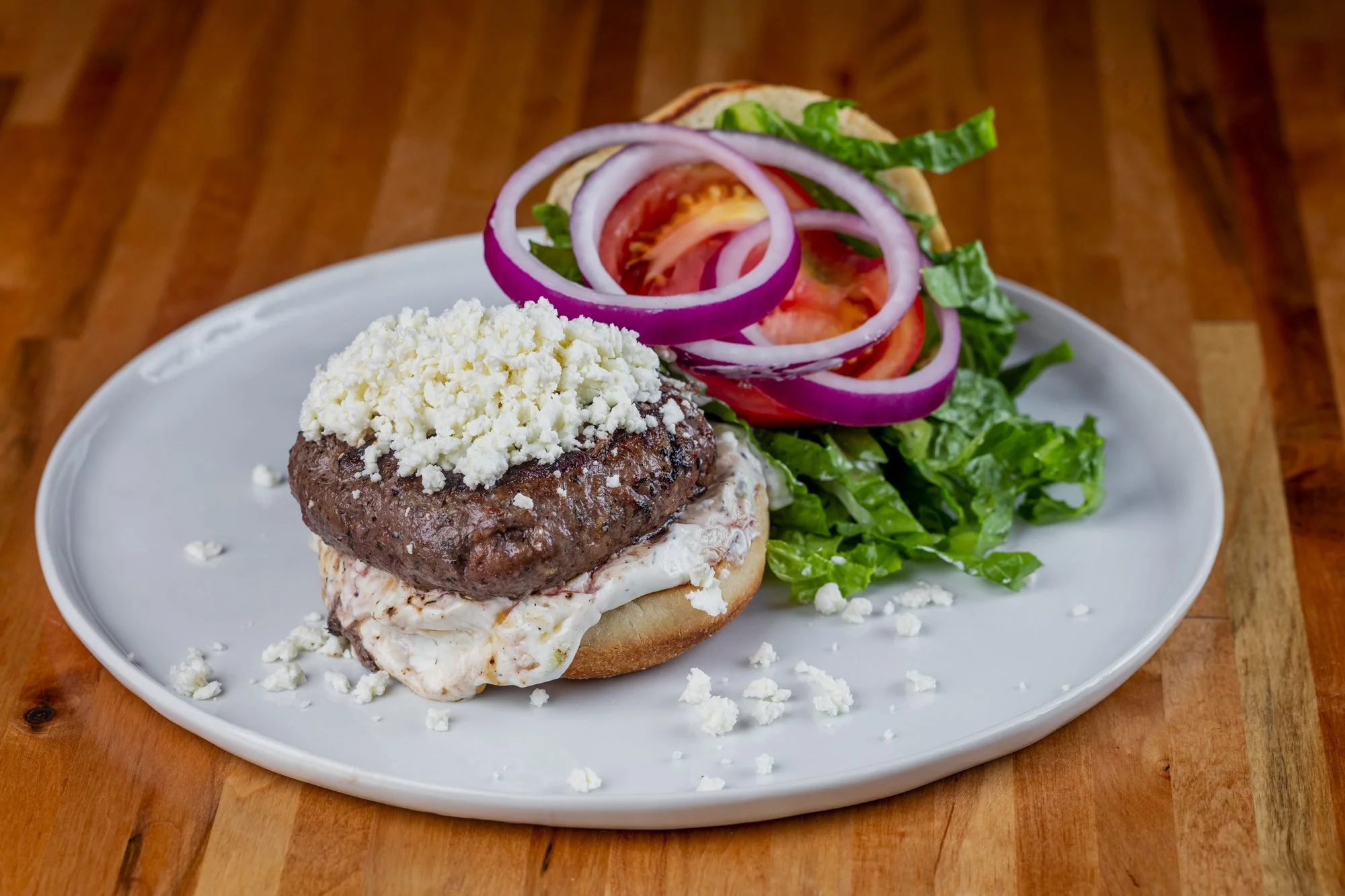 A plate with a hamburger containing a beef patty, feta cheese crumbles, and tzatziki sauce, with lettuce, tomato, and red onion slices on a wooden surface.