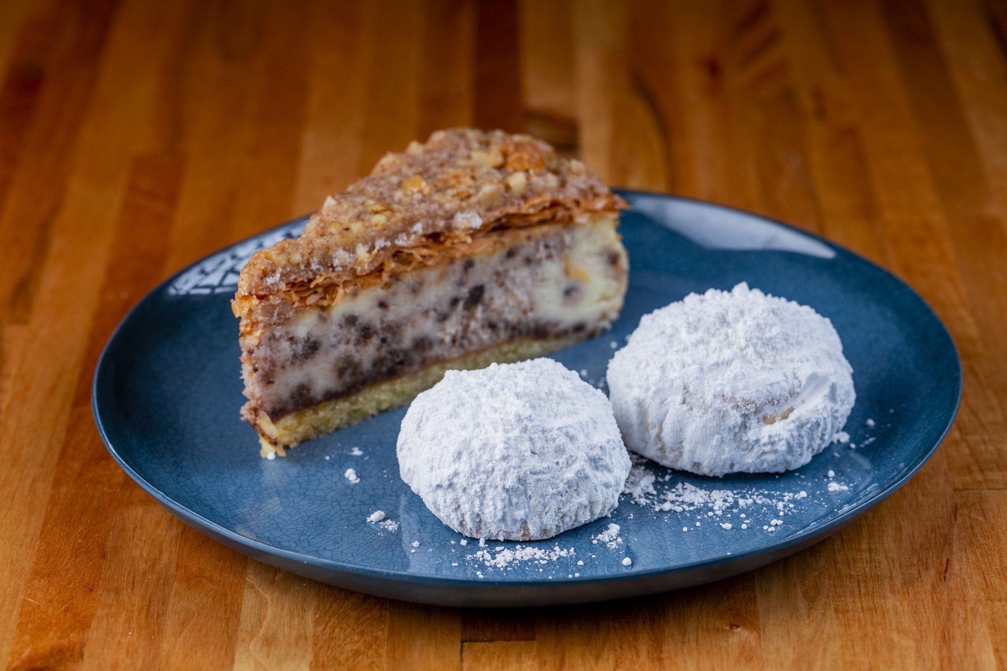 Slice of baklava cheesecake on a blue plate with two powdered sugar-coated cookies, on a wooden surface.