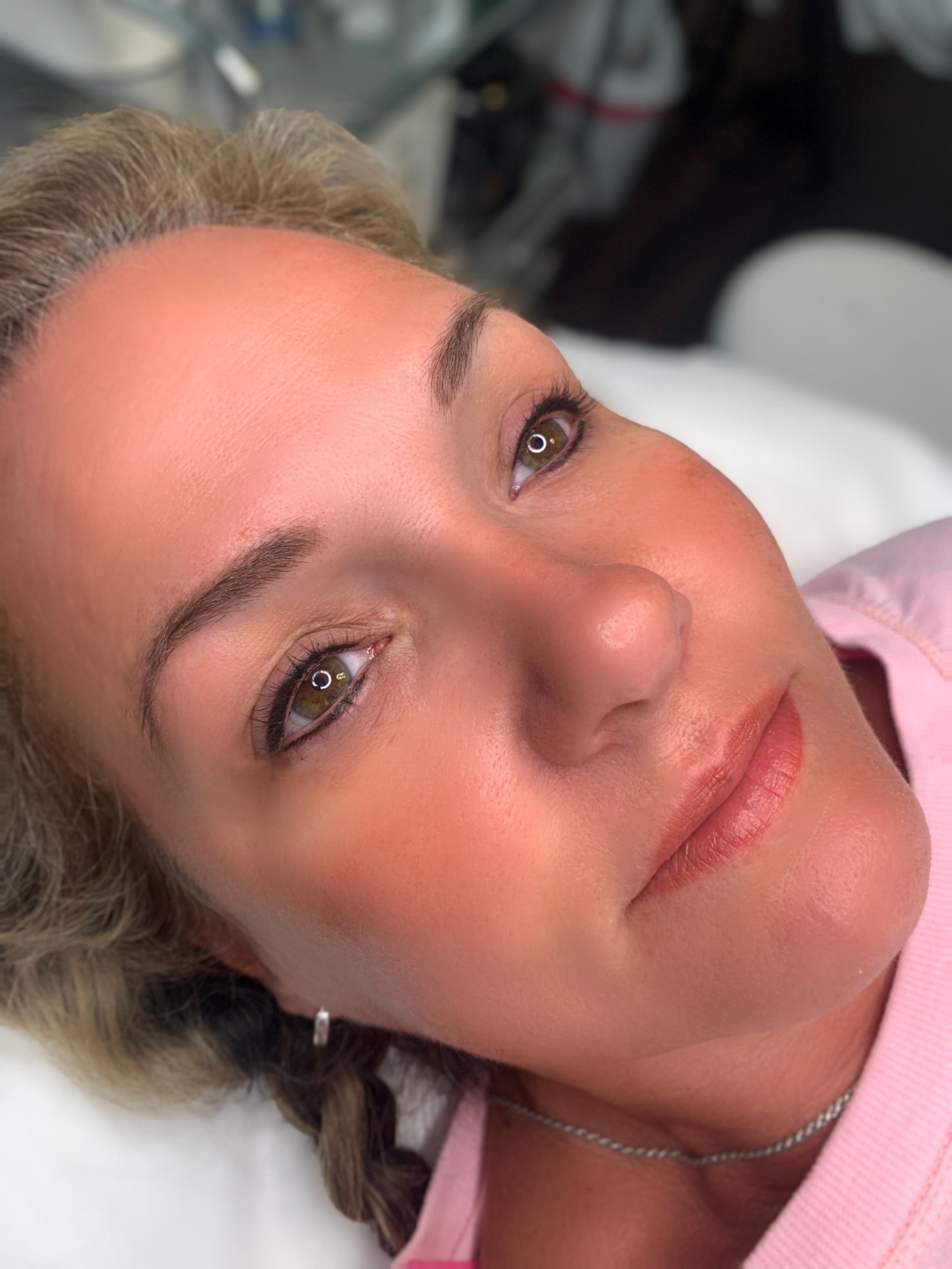 A woman with light brown hair lying down, looking up with hazel eyes, wearing a pink top and a silver necklace, with a neutral expression.