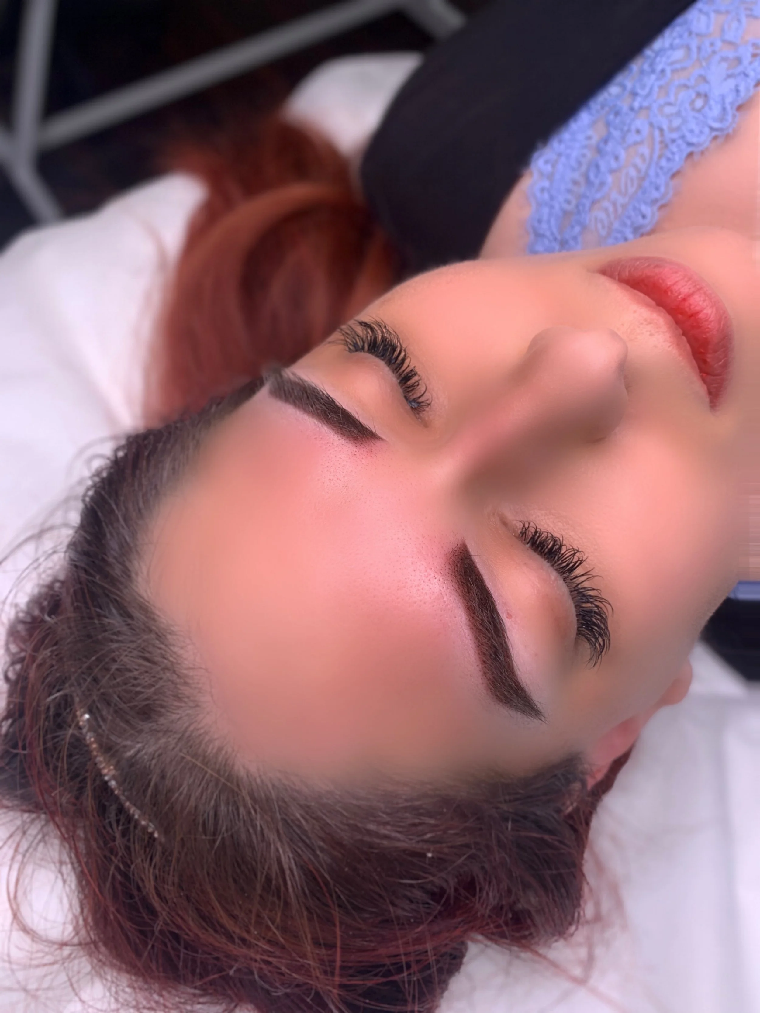 A woman with reddish-brown hair lying down with eyes closed, showcasing curled eyelashes and well-groomed eyebrows.