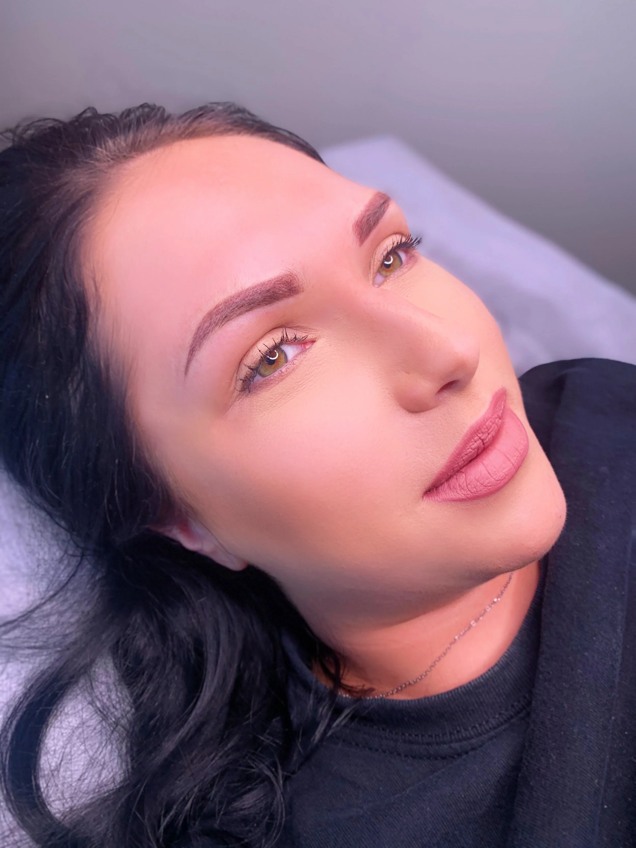 Close-up of a woman lying down with newly done eyebrows and long eyelashes, dark hair, and wearing a black shirt.