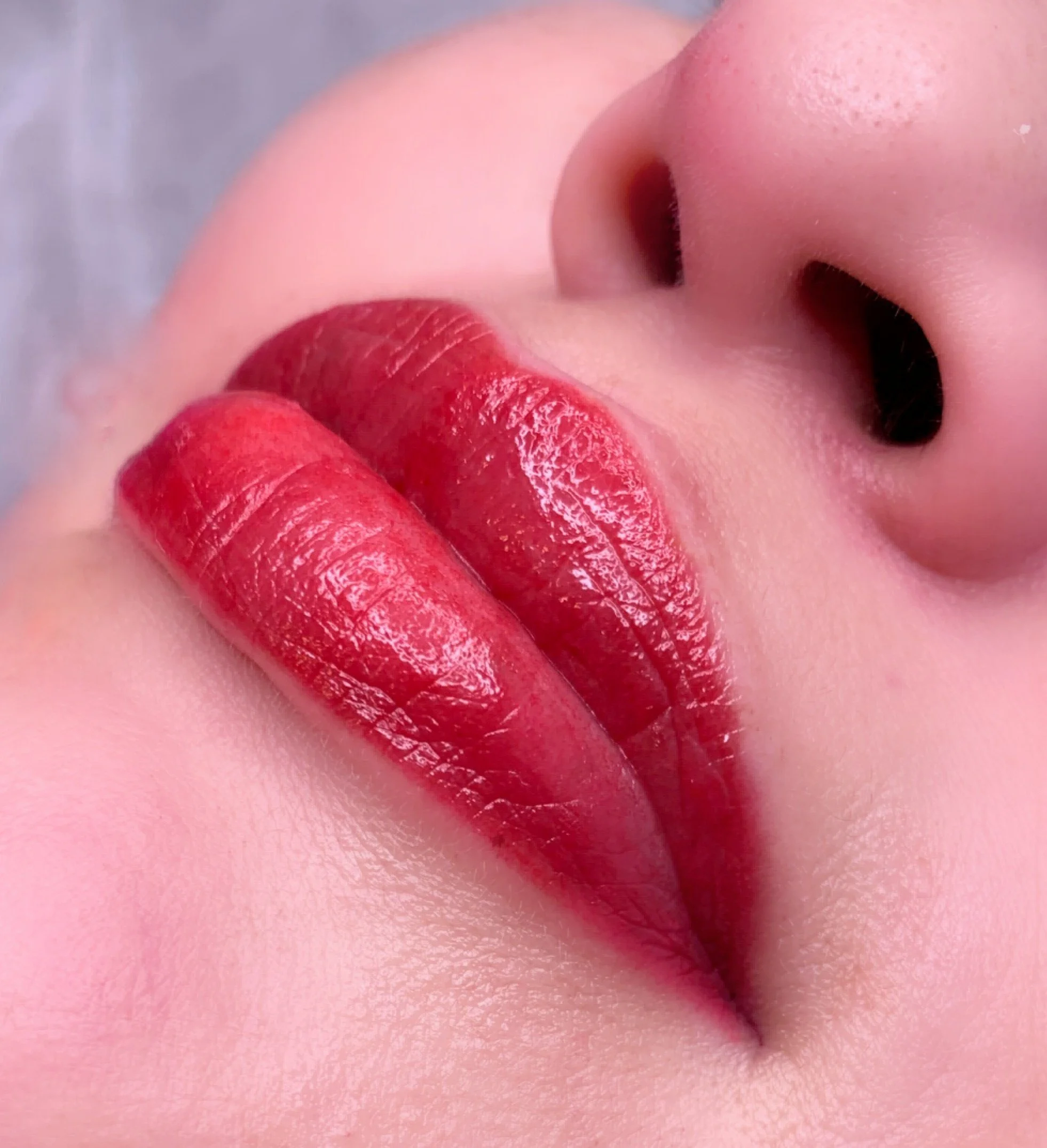Close-up of a person's face with bright red lipstick, showing lips, nose, and part of the cheek, with a soft pink background.