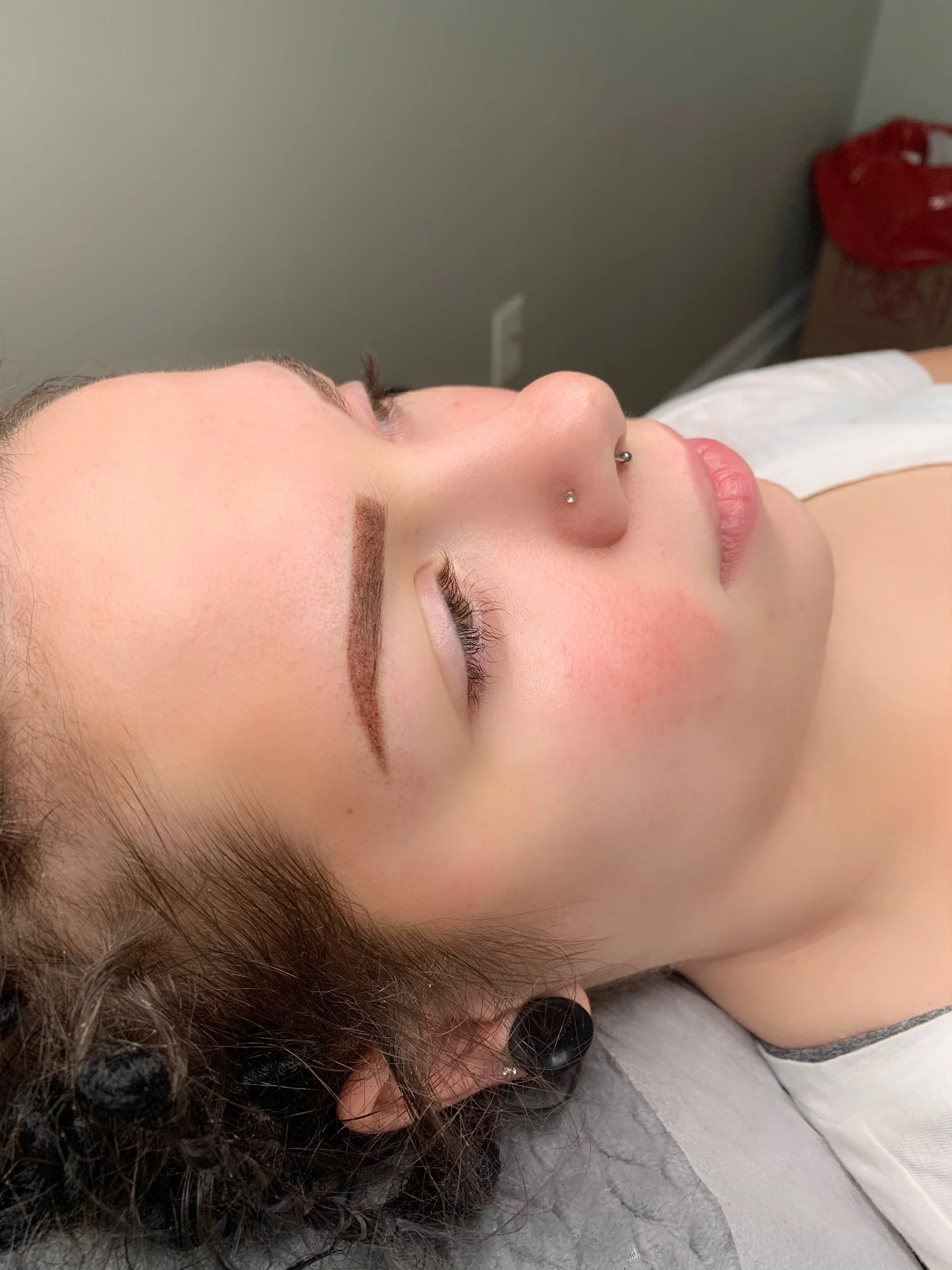 Close-up of a woman's face lying down on a table, with visible eyebrows, nose piercings, and closed eyes, in a medical or beauty setting.