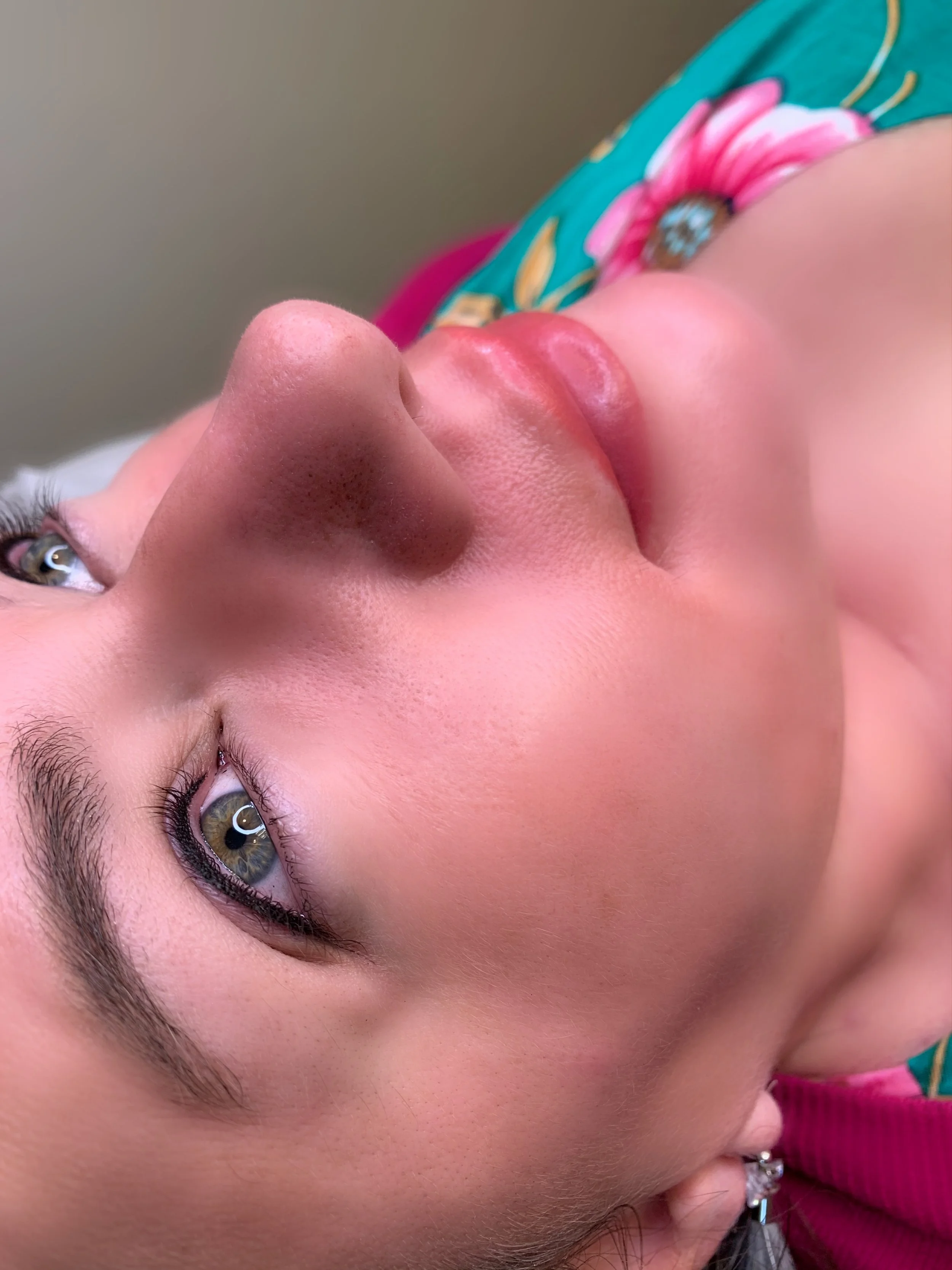 Close-up of a person's face lying on their side, showing blue-green eyes, light skin with pink blush, pink lips, and arched eyebrows. A floral clothing pattern is partially visible in the background.