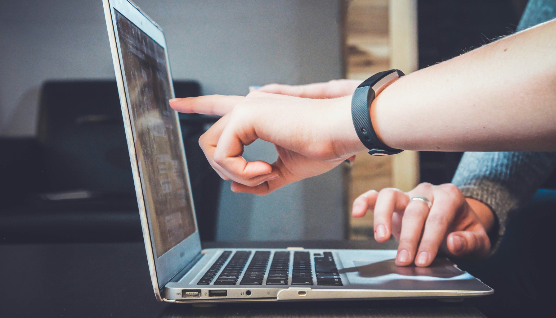A person pointing at a laptop screen with another person's hand resting nearby.
