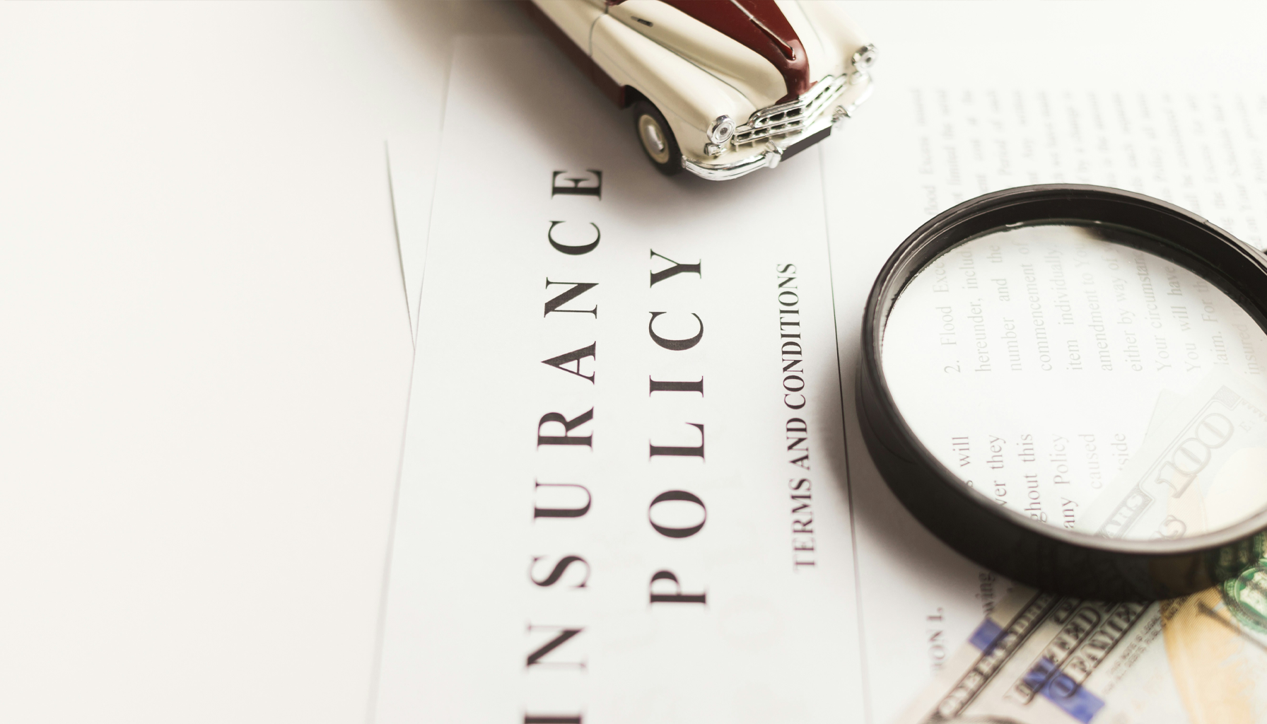 A legal document titled 'Insurance Policy' titled on a table. A toy vintage car, a magnifying glass, and a hundred-dollar bill are placed on or near the document.