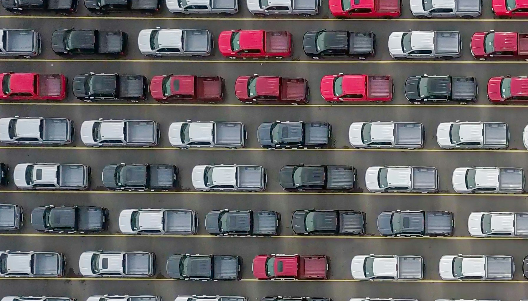 Aerial view of multiple parked pickup trucks in a parking lot, arranged in neat rows with yellow lines marking spaces. The trucks are mainly white, black, gray, with some red vehicles interspersed.