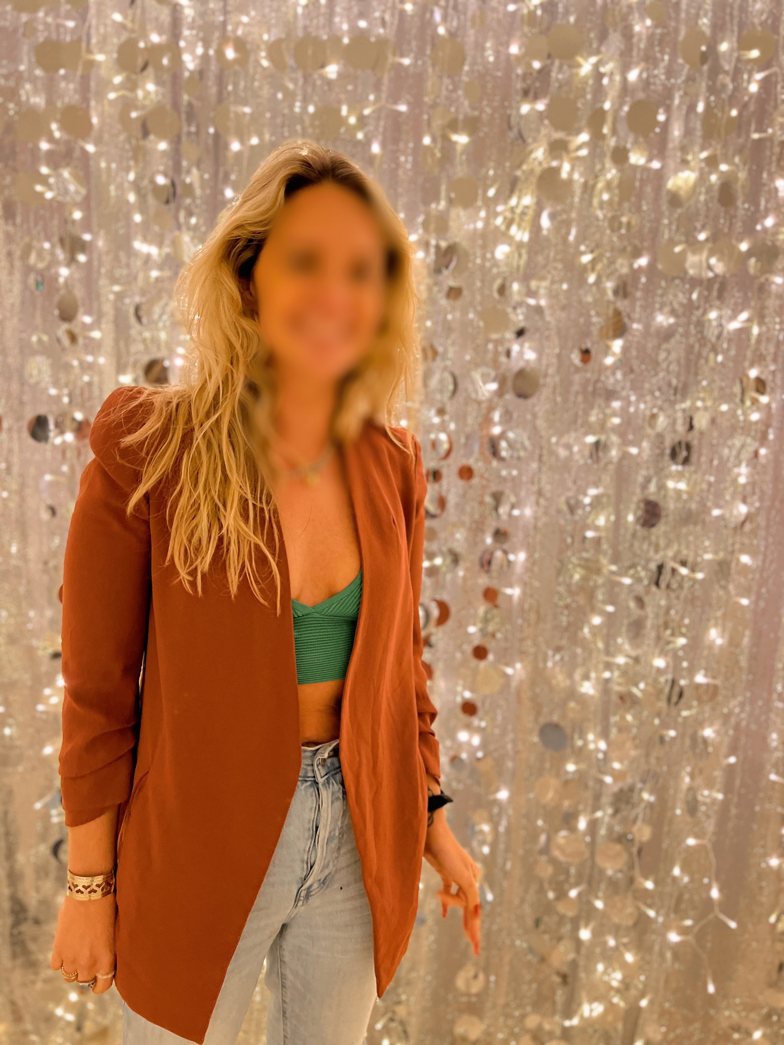 A woman with blonde curly hair, blurred face, wearing a rust-colored blazer, green crop top, and light-wash jeans, standing in front of a shimmering, sequin-covered curtain backdrop.