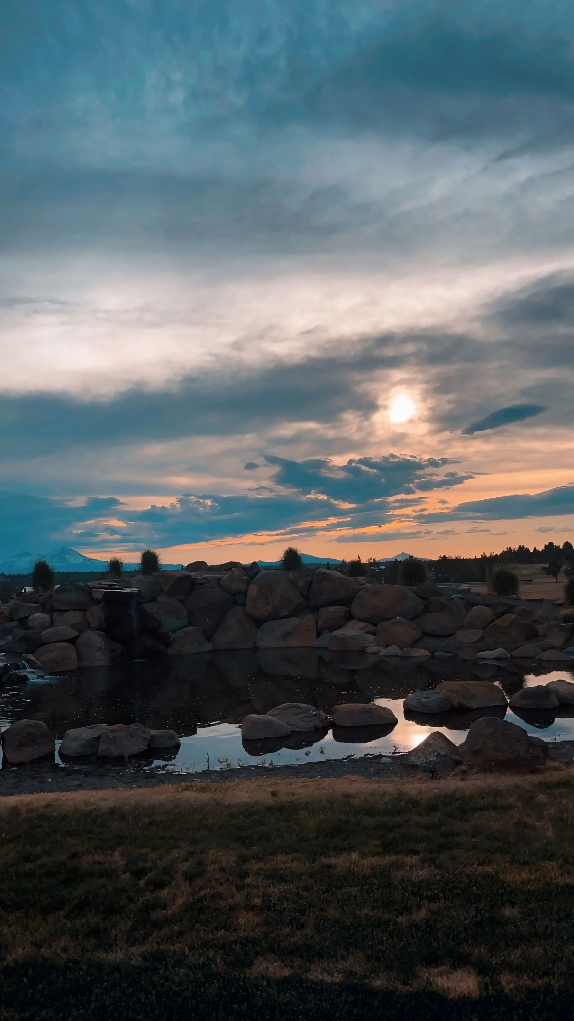 A landscape at sunset with a cloudy sky, a small pond, and rocks along the shore.