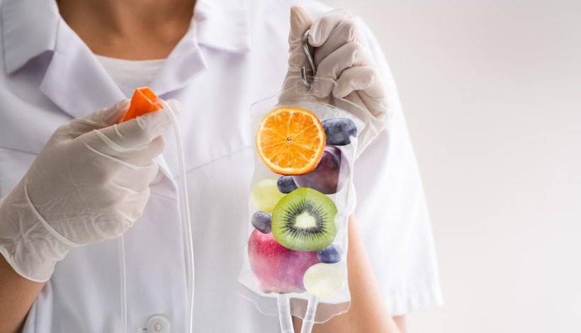 A person in a white lab coat and gloves holds a beaker with sliced orange, blueberry, kiwi, grape, apple, and banana inside, with a syringe in the other hand.