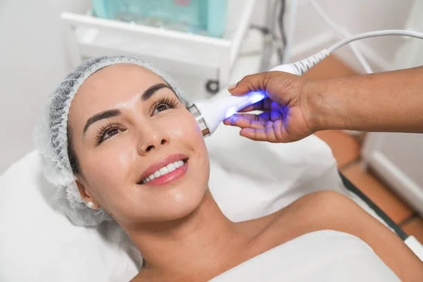 A woman receiving a cosmetic facial treatment using a handheld device emitting blue light, while lying in a clinical setting.