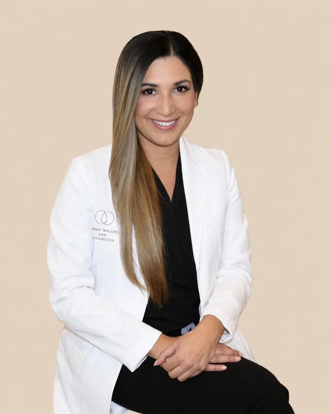 A woman with long hair, wearing a white medical coat and black attire underneath, sitting against a beige background, smiling at the camera.