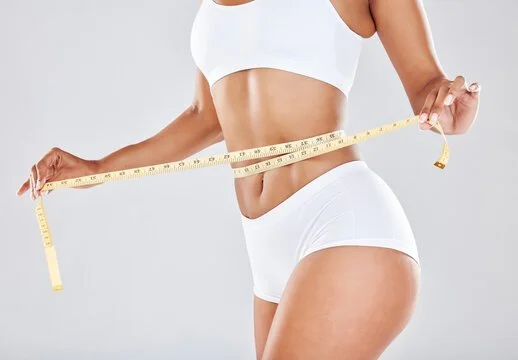 A woman in white workout clothes measuring her waist with a yellow tape measure.