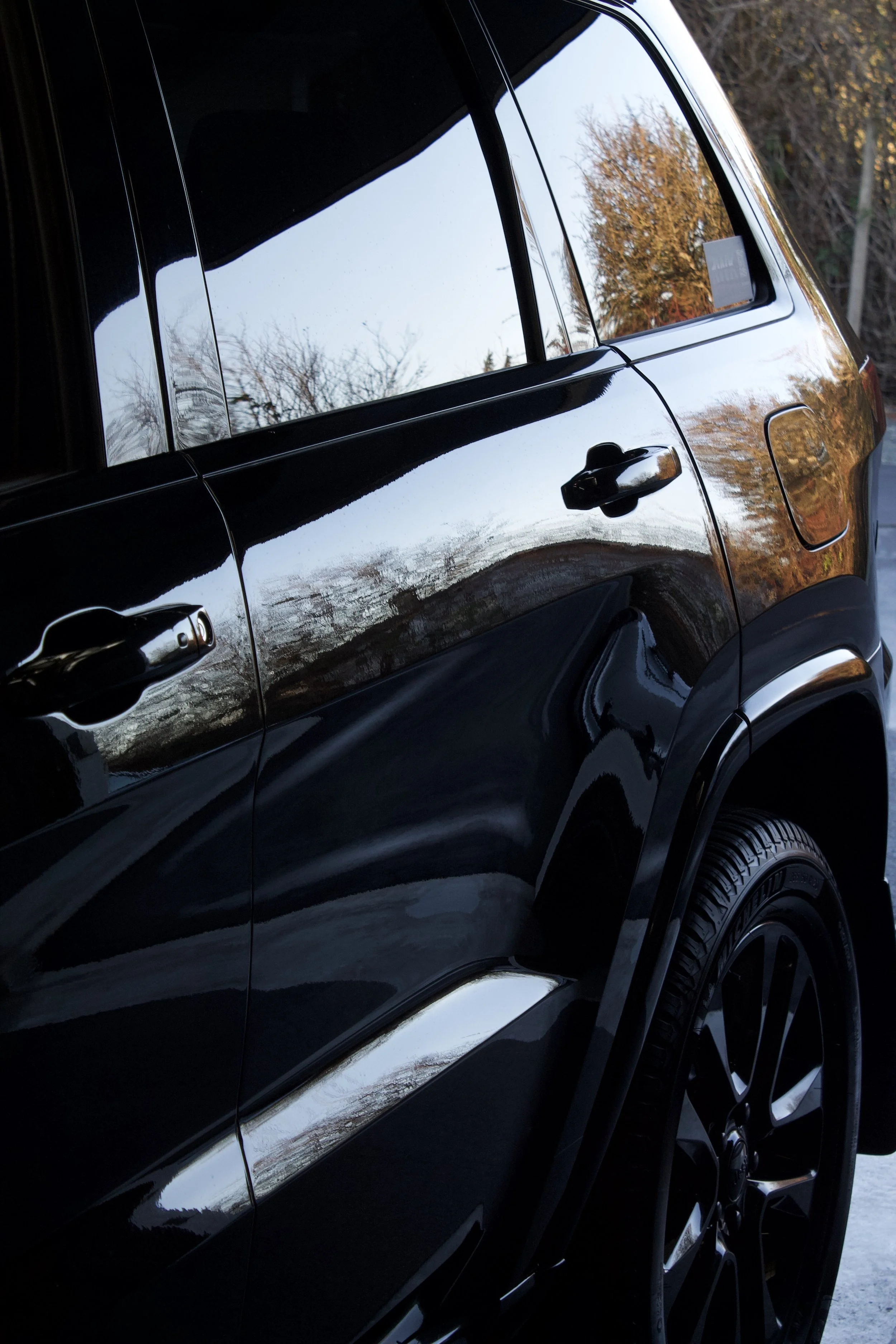Close-up of a shiny black car with reflections of trees and sky on its surface, showing the rear passenger side.