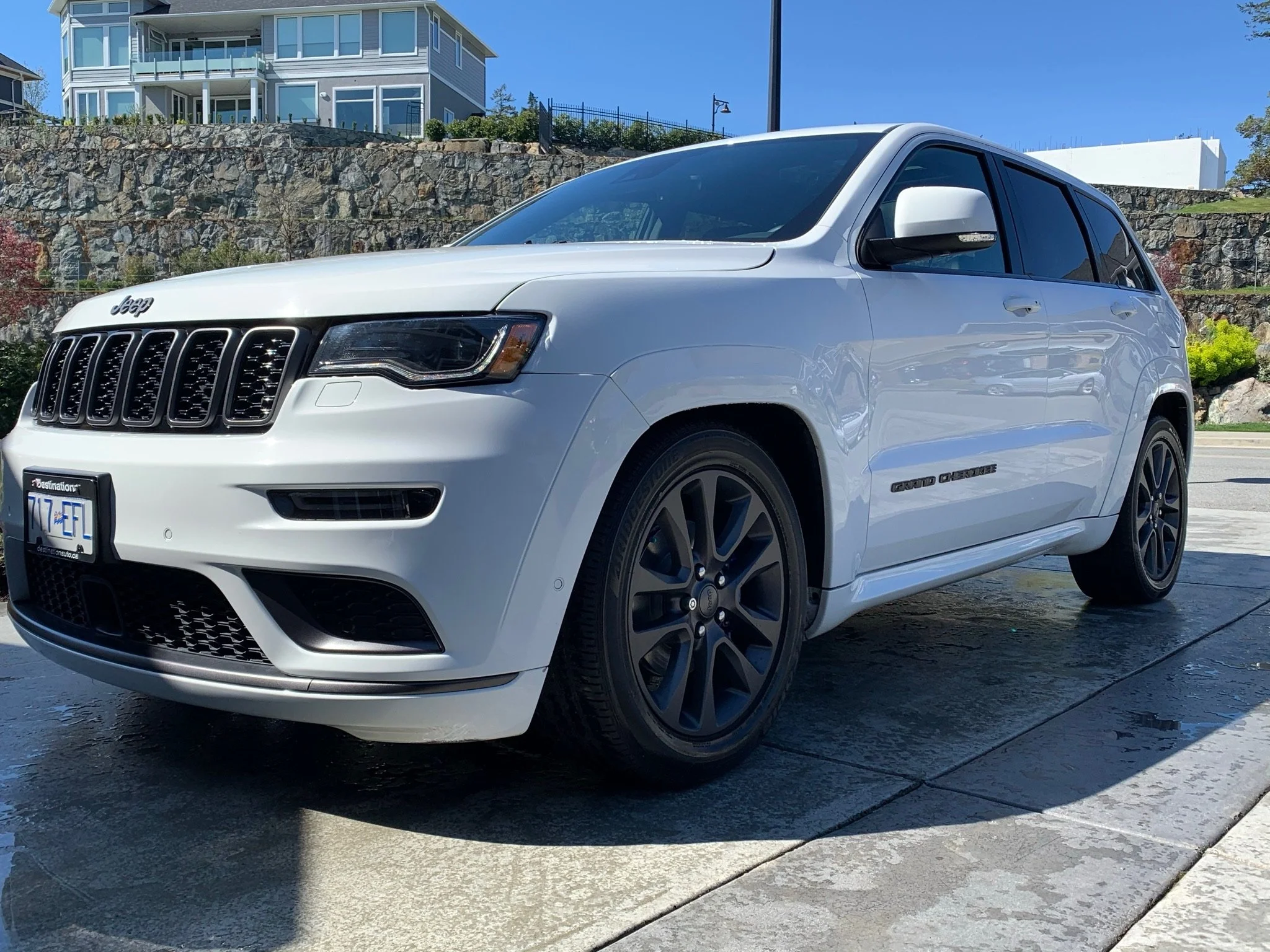 White Jeep Grand Cherokee freshly washed and detailed parked on a driveway with a stone retaining wall and a multi-story house in the background.