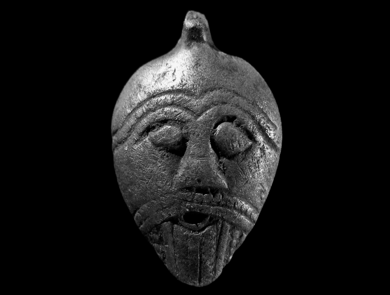Fotografía de un pendiente de plata en forma de máscara facial del período de Vendel, hallado en Selandia, Dinamarca. Se observa el desgaste natural del metal por uso antiguo.