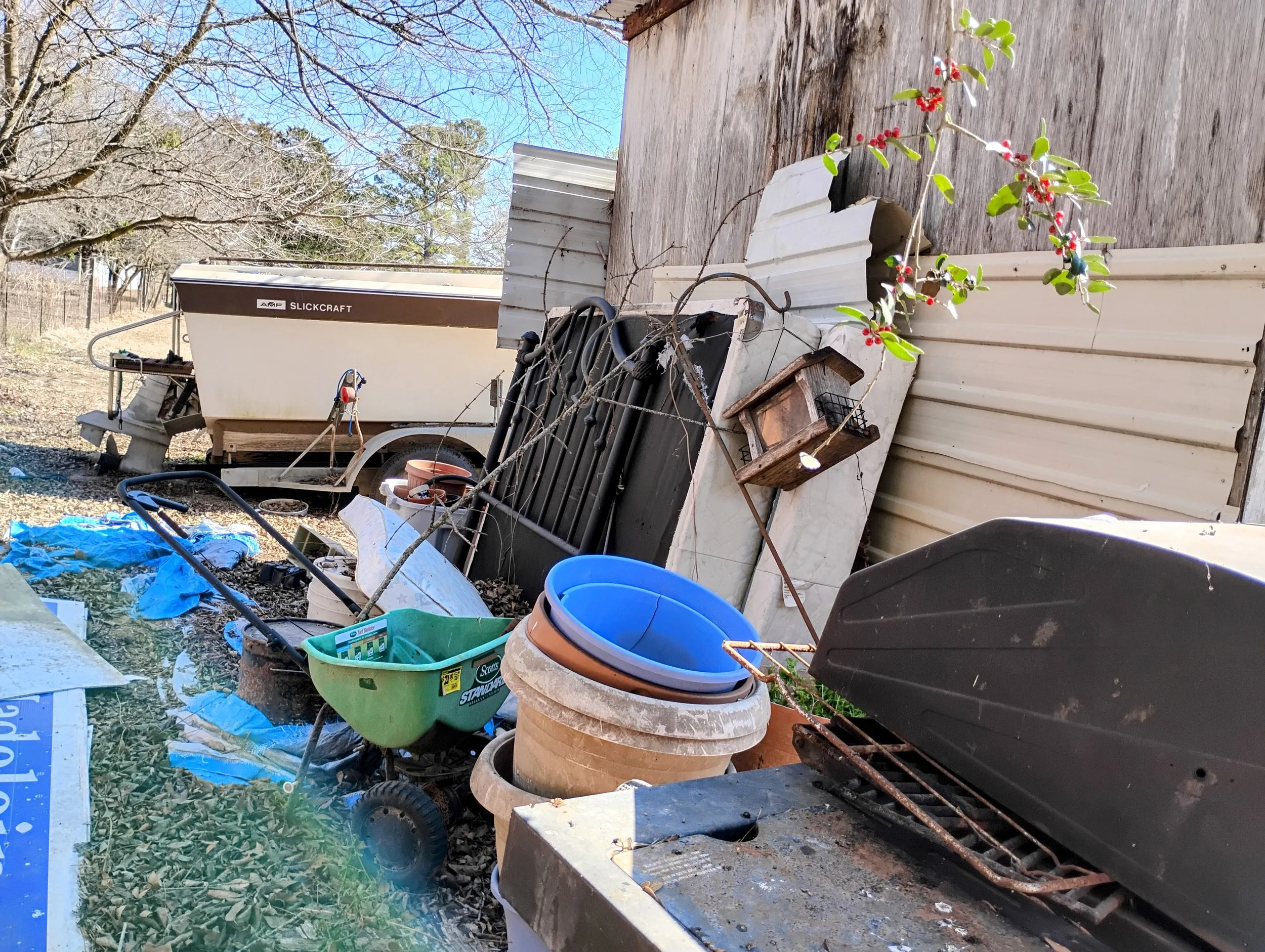 An old boat. wheelbarrow, planters, and trash outside a wooden building.