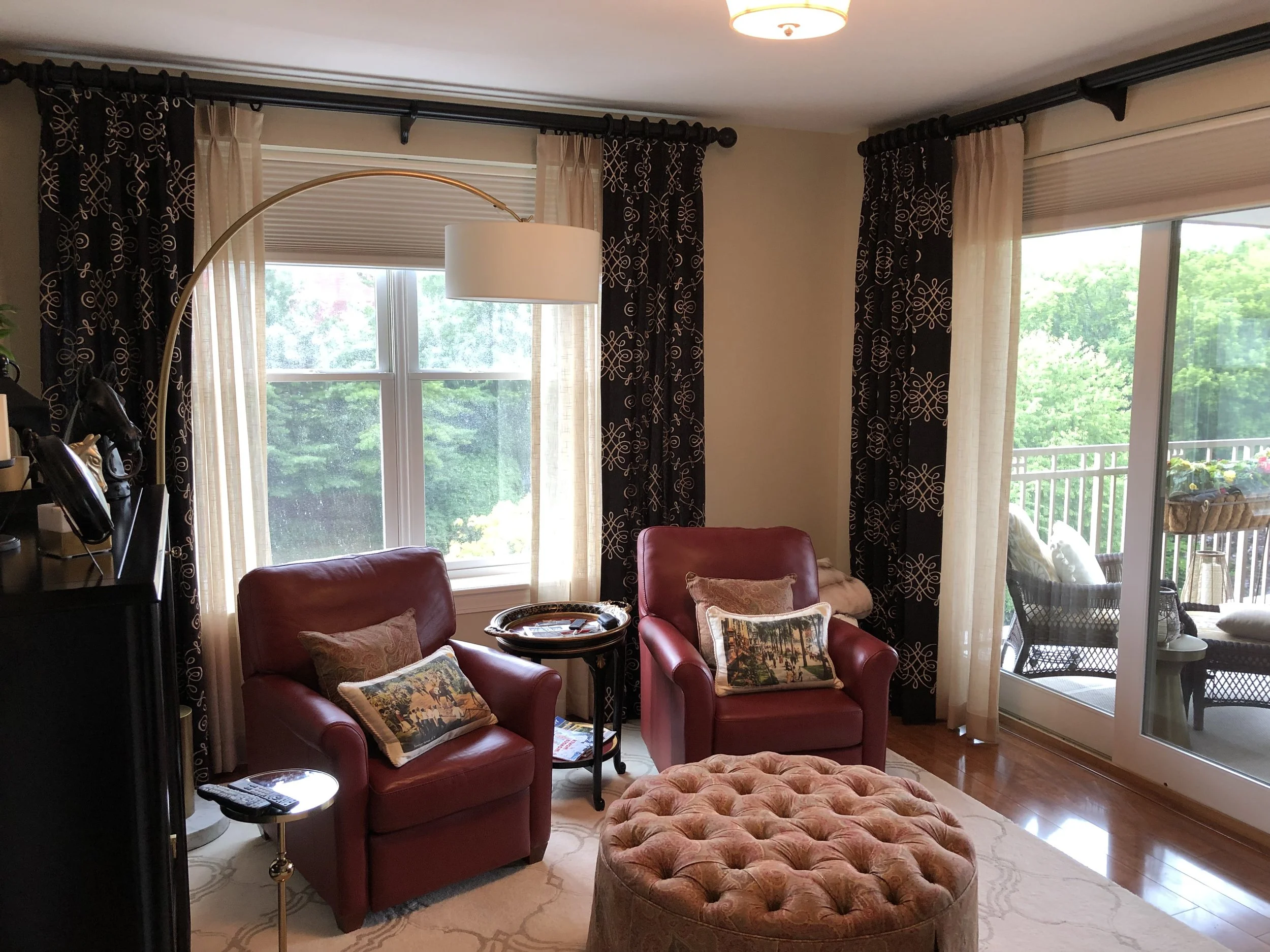 Living room with two red armchairs, a tufted ottoman, black and white patterned curtains, and a sliding glass door leading to a balcony with outdoor furniture and potted plants.