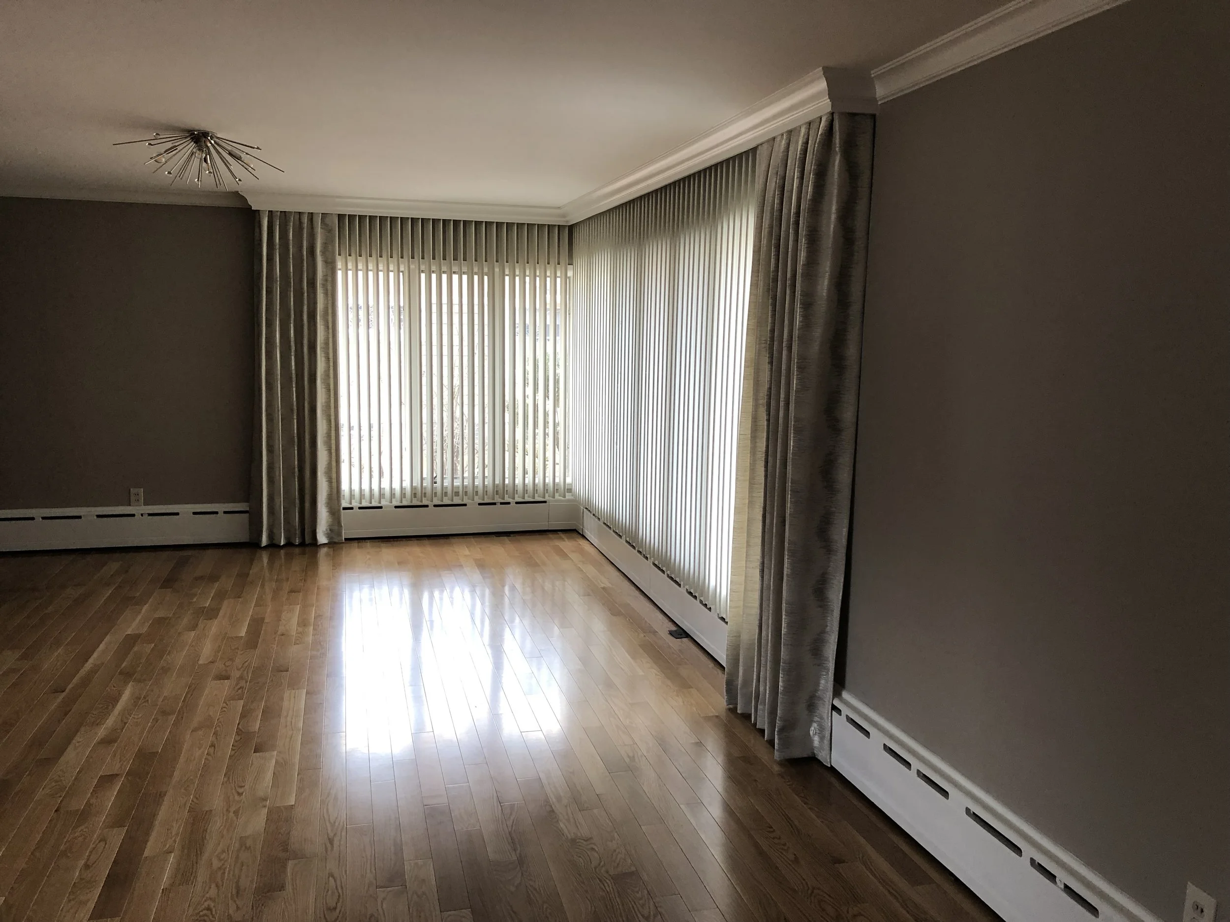 Empty living room with hardwood floors, large windows with vertical blinds, gray curtains, and a modern ceiling light fixture.